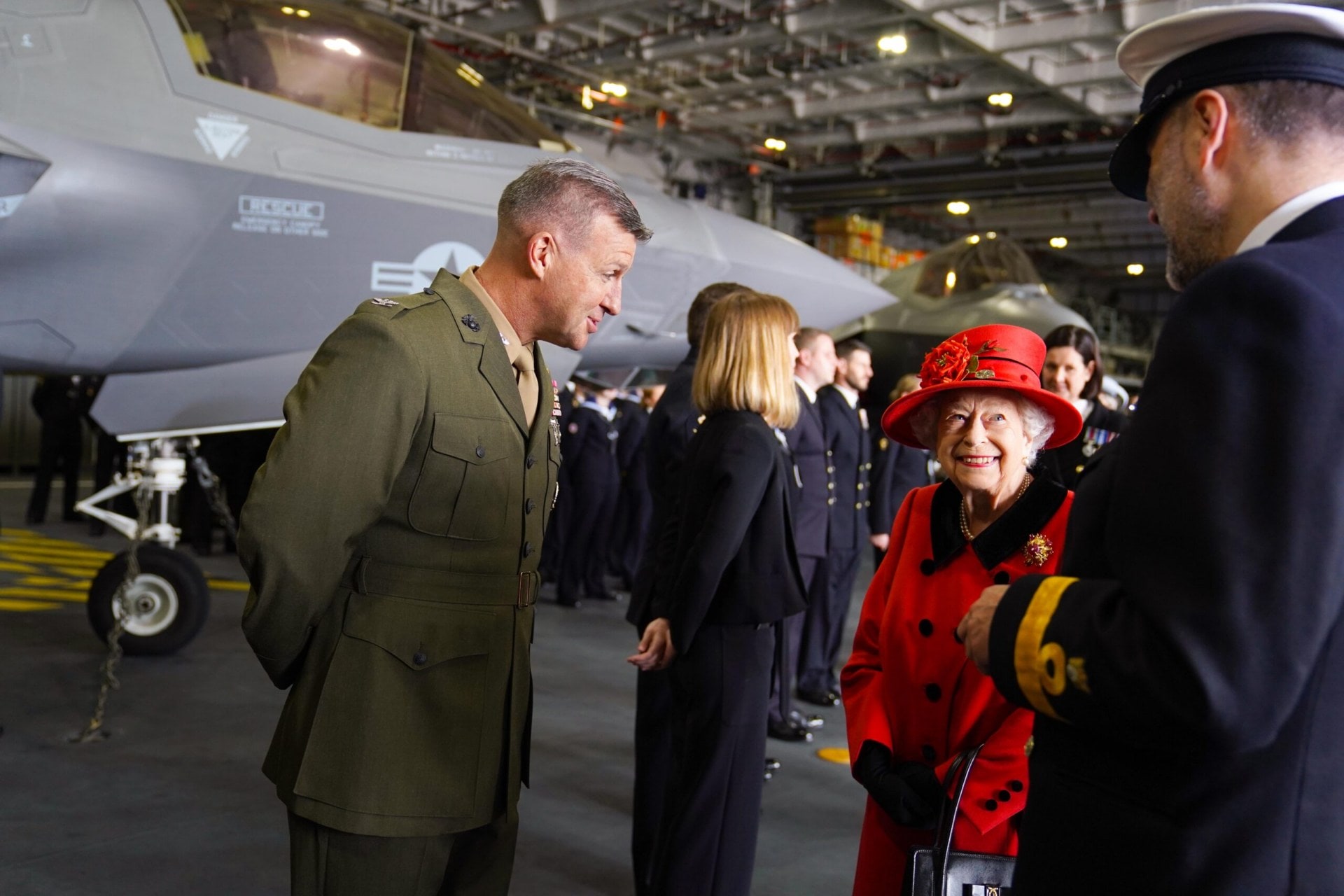 Queen Elizabeth II wearing a red coat and speaking with two of the crew of the HMS Queen Elizabeth