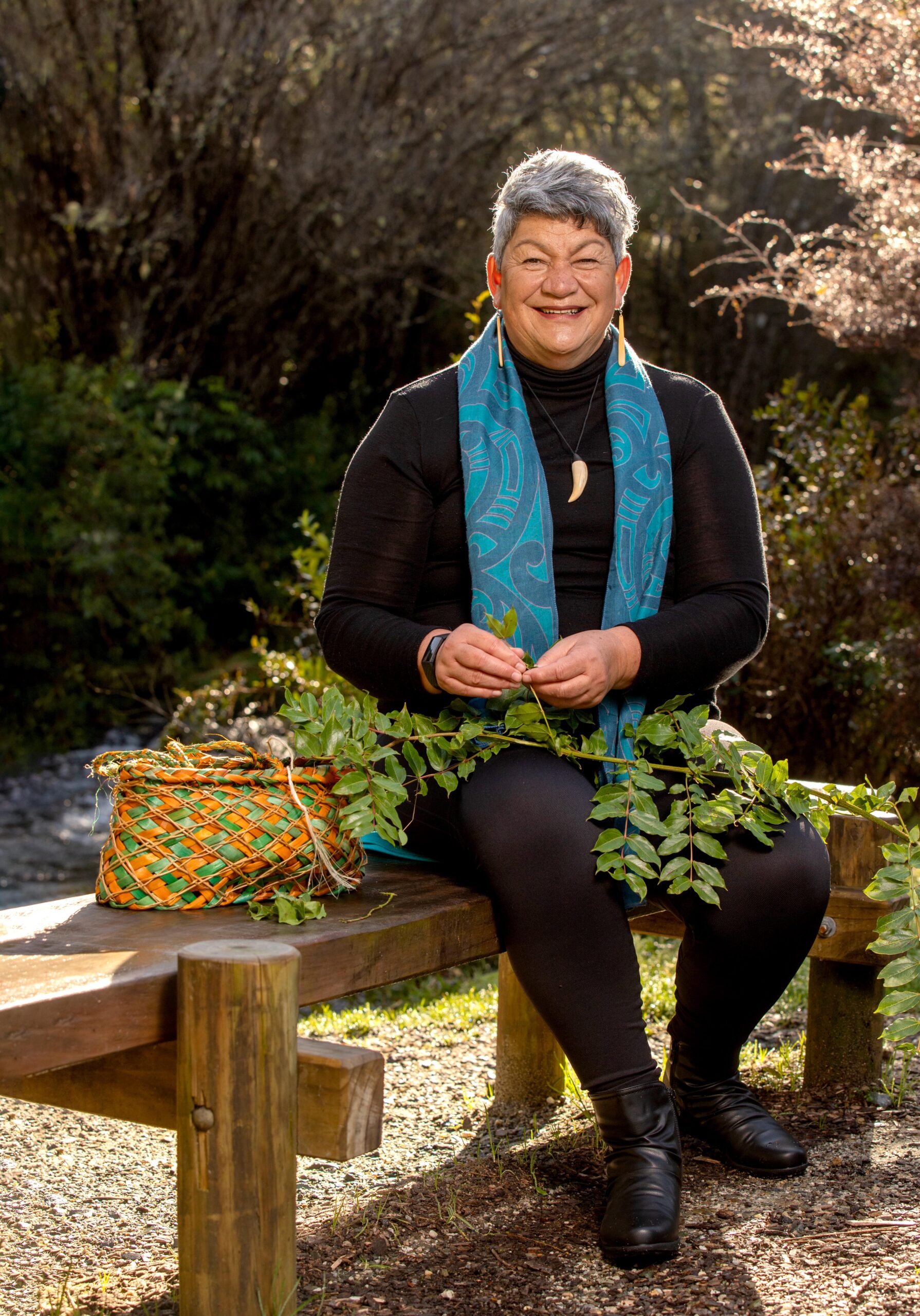 Donna Kerridge sitting on a wood garden bench holding a green plant and wearing a black outfit and blue scarf