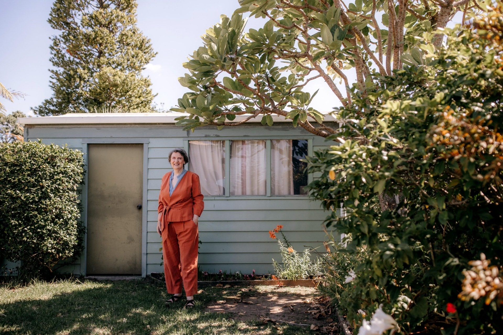 Helen Clark standing outside garden shed wearing coral red outfit