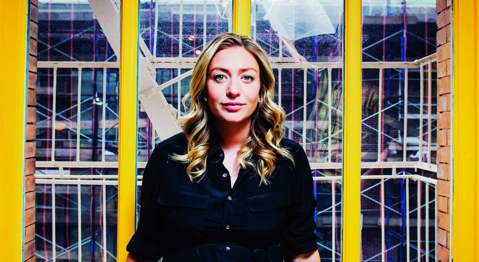 Whitney Wolfe Herd wearing black blouse leaning against a bright yellow window ledge in the Bumble offices 