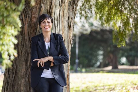 Rachel Taulelei leaning against a tree wearing a black suit and greenstone necklace