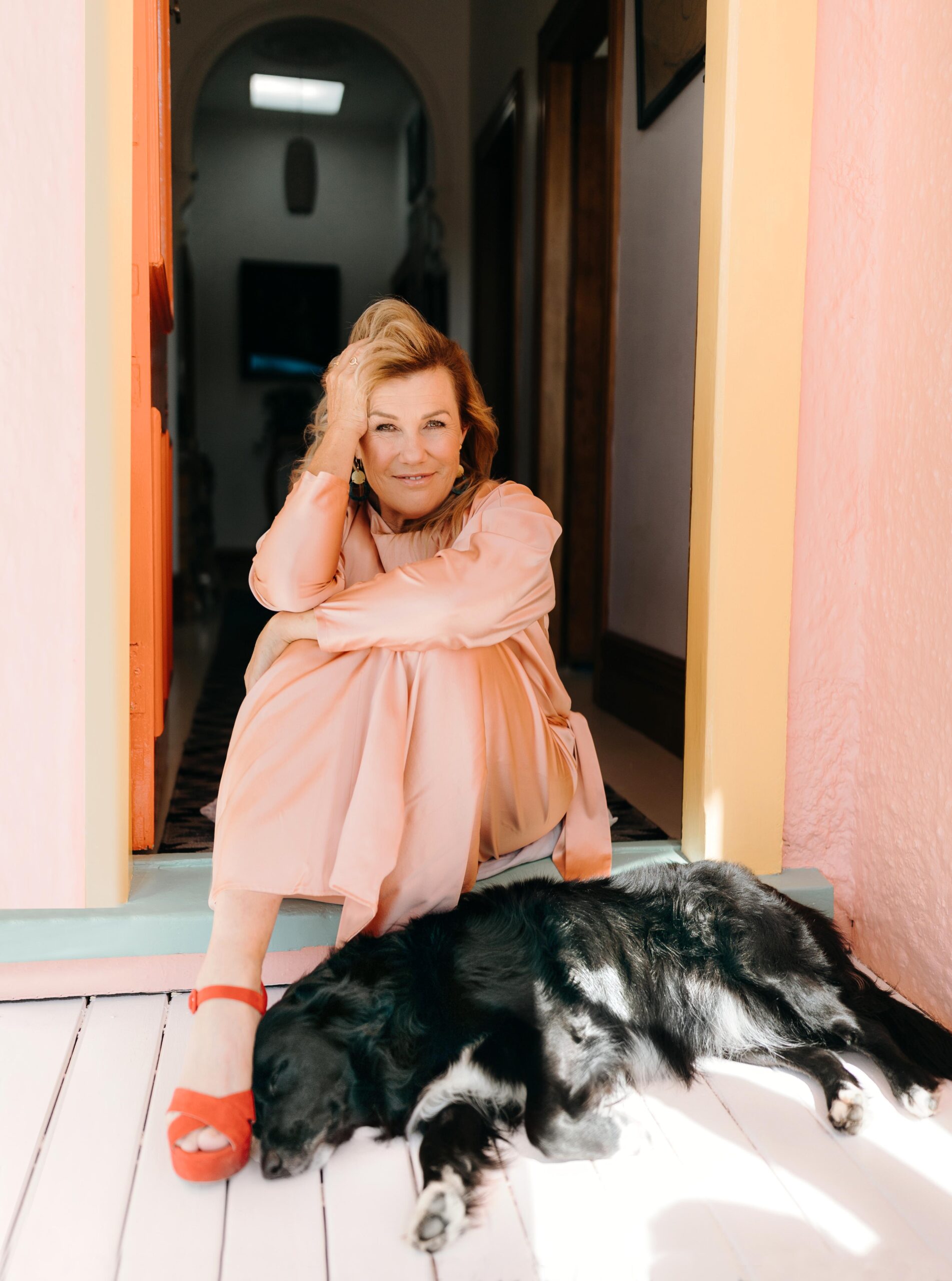 Robyn Malcolm sitting in doorway next to her pet dog. She's wearing a pink dress and red shoes.