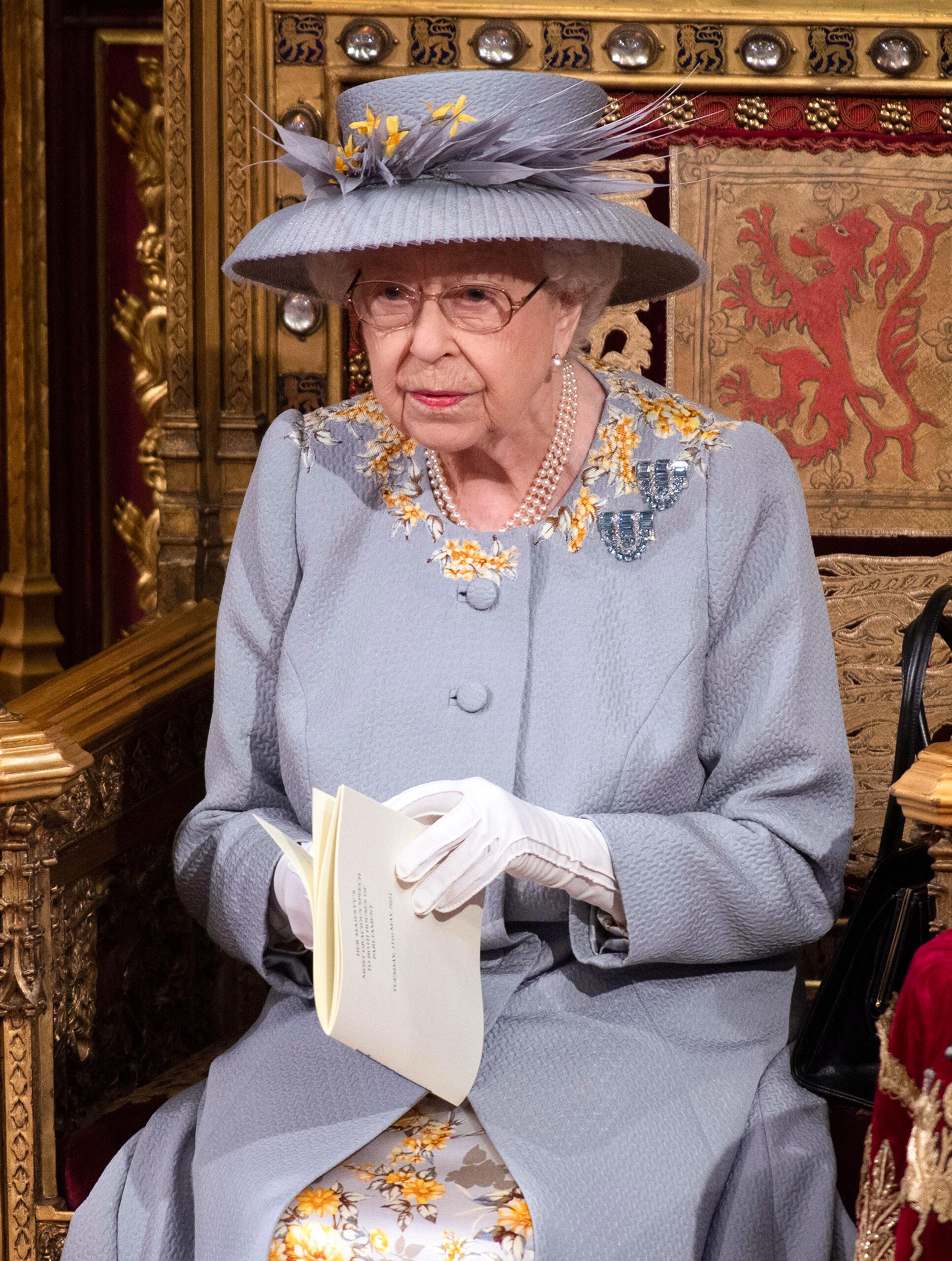 Queen Elizabeth II wearing light blue coat with yellow floral detailing and matching hat sitting on a gold and red chair at the State Opening of Parliament