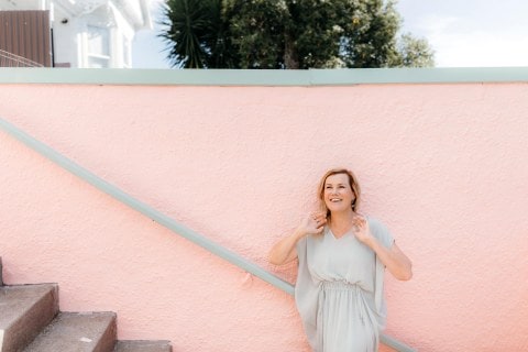 Robyn Malcolm wearing light blue dress laughing while standing against pink background