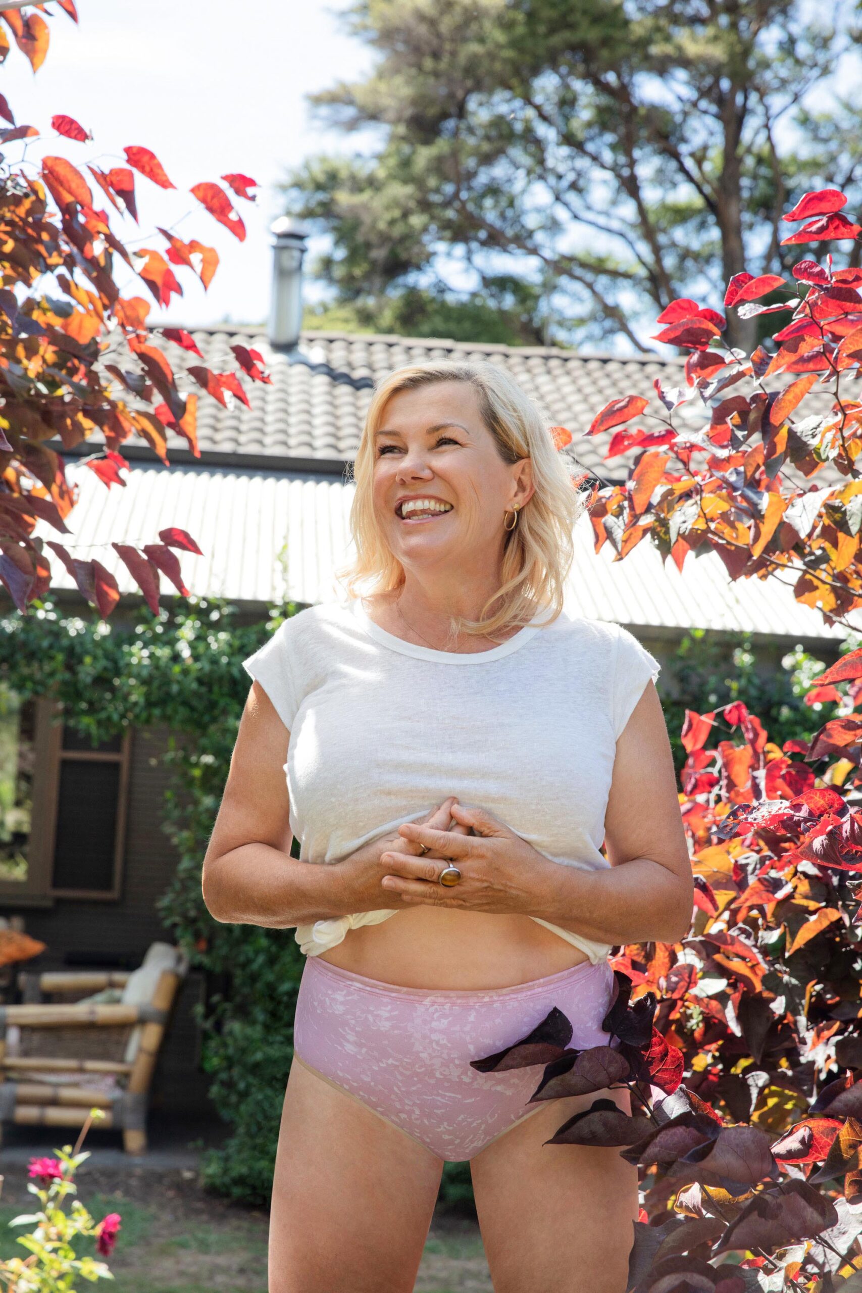 Robyn Malcolm laughing while standing in garden wearing pink Robyn's Undies and a white t-shirt