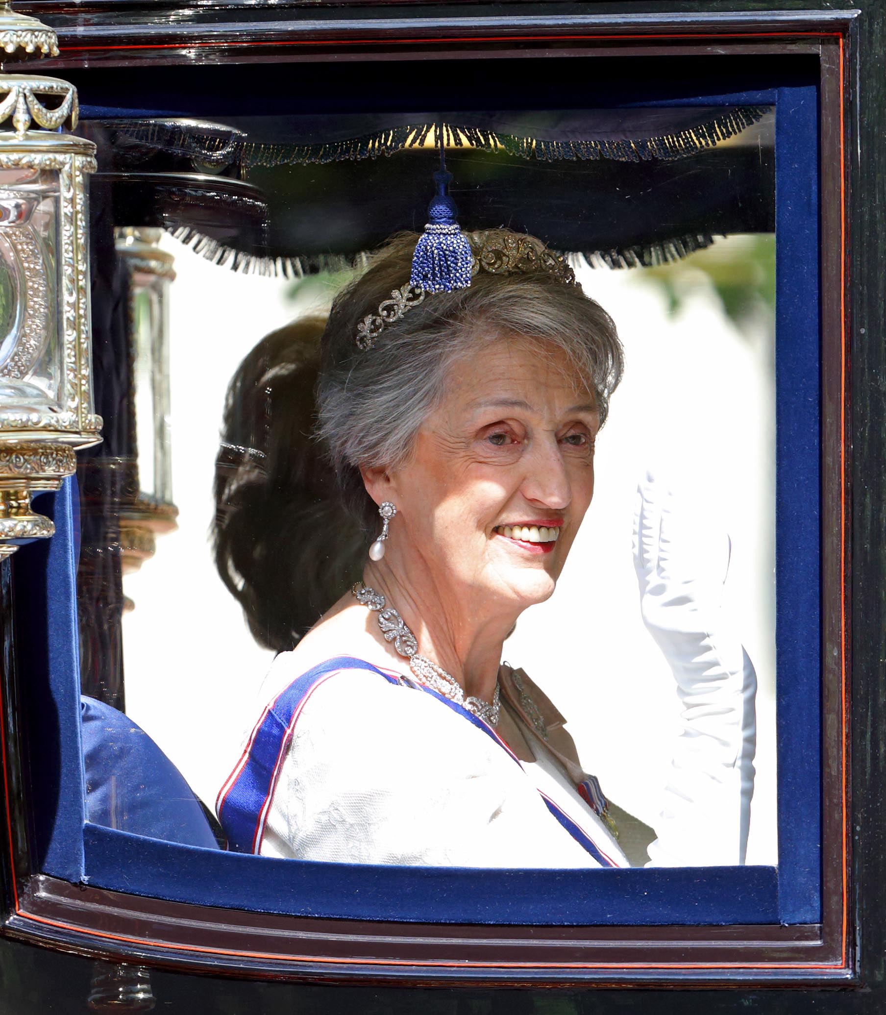 Lady Susan Hussey waving and smiling from a carriage. She is wearing diamond jewellery, a white glove and a blue sache