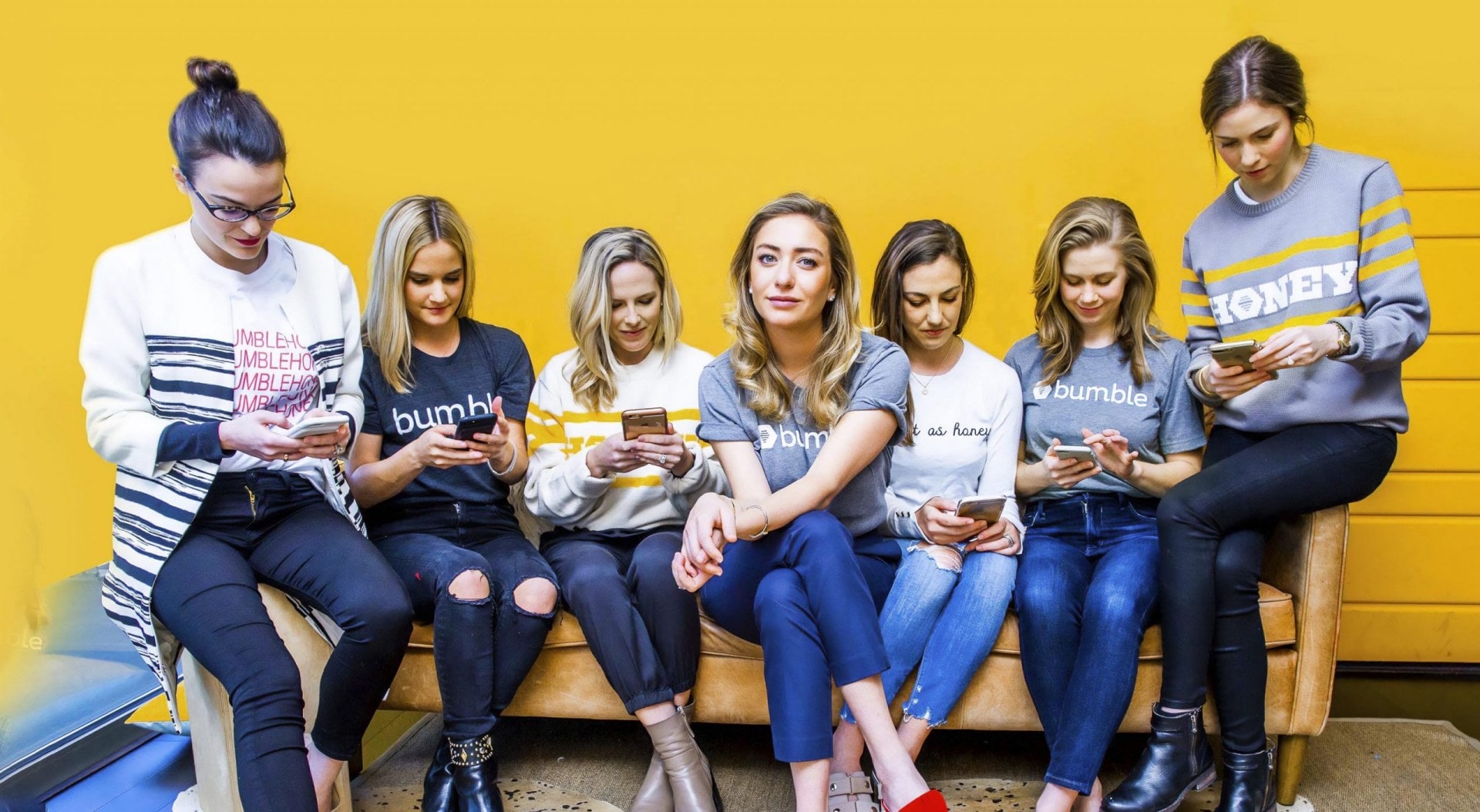 Whitley Wolfe Herd sitting on an orange couch with six women looking down at their mobile phones