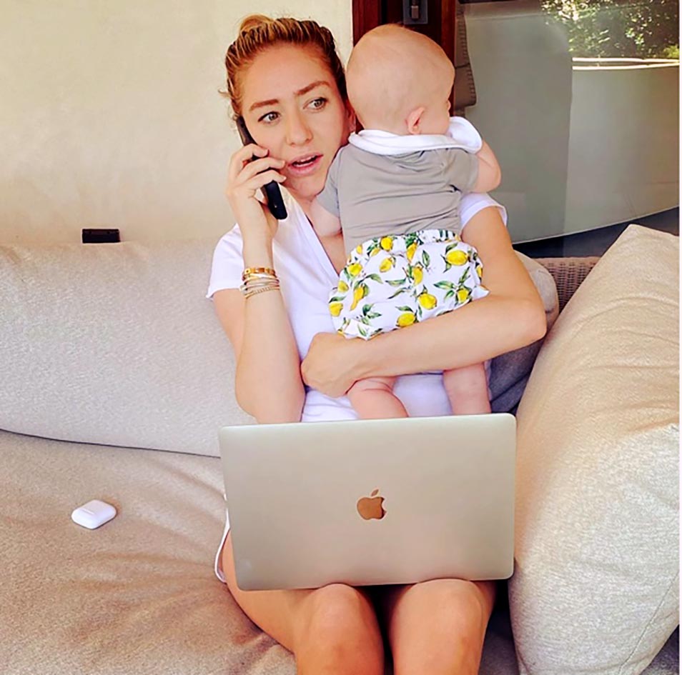 Whitney Wolfe Herd sitting on a couch holding her baby Bobby with one hand while speaking on a mobile phone she's holding on the other
