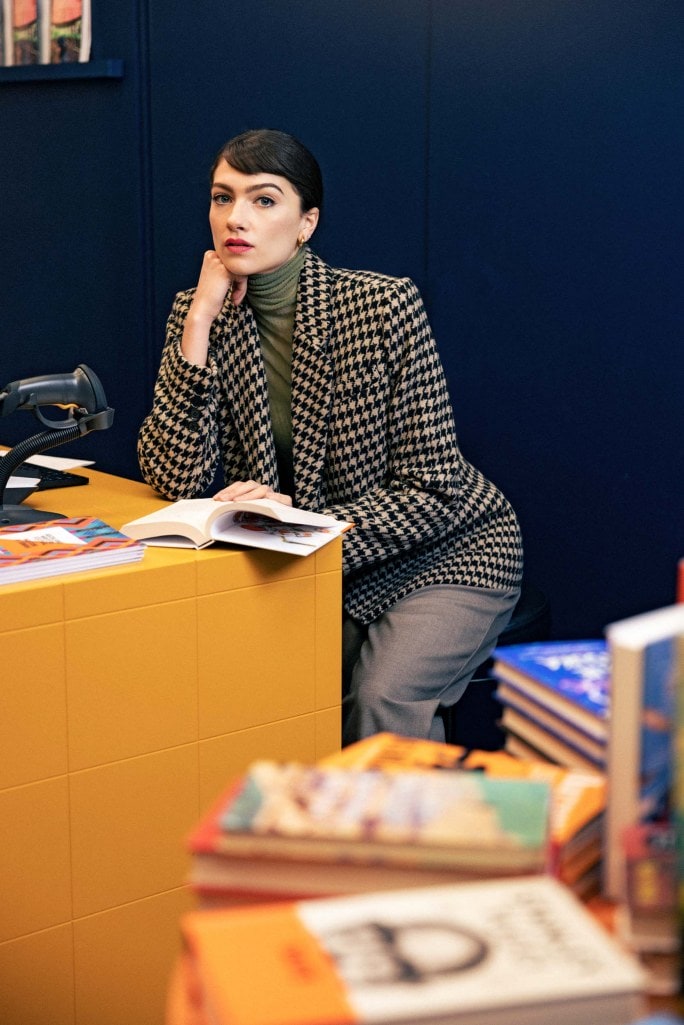 a woman wearing a patterned blazer sitting with stacks of books