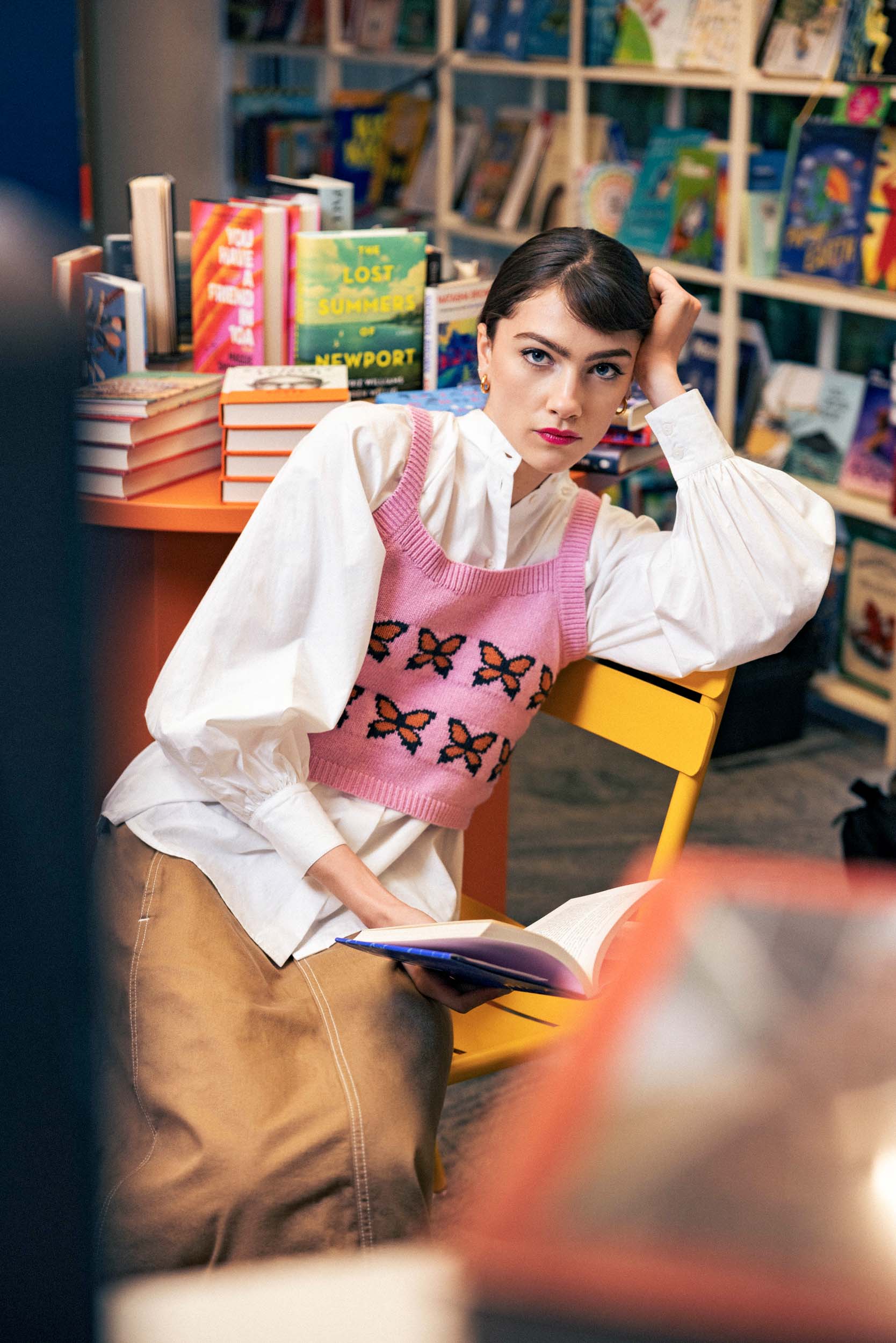 a model wearing a pink knitted vest and white shirt sitting in a bookstore