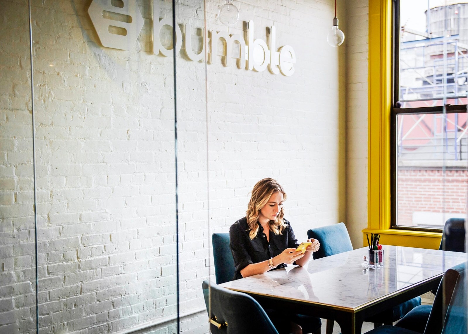 Whitney Wolfe Herd sitting in an office with white brick walls and a Bumble logo