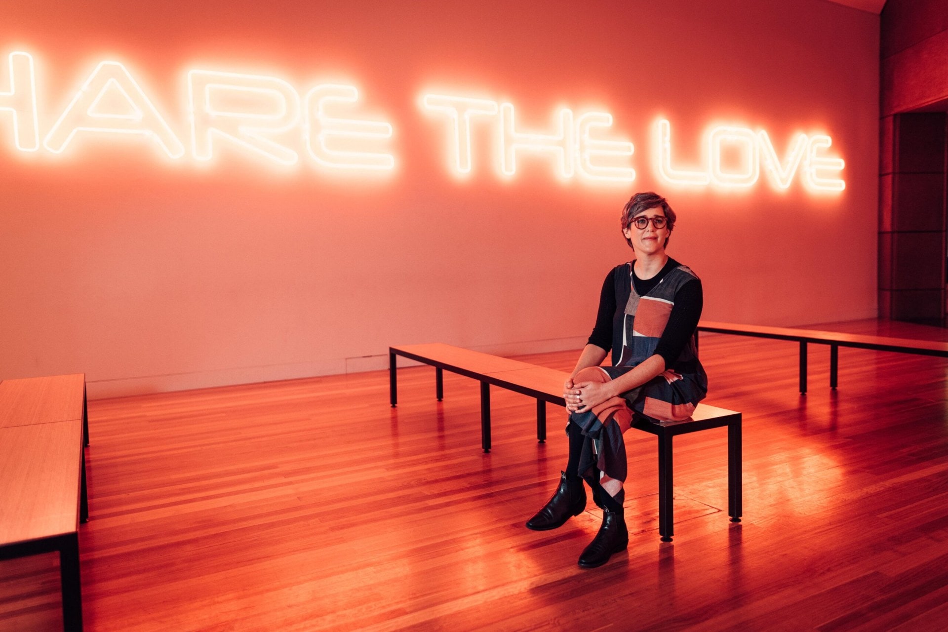 Lizzie Bisley sitting on a bench in a room with neon writing on the wall saying Share the Love