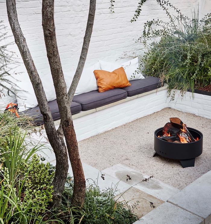 Sunken garden among a white brick wall, with built in bench seat with grey and white cushions, paving stones and gravel are on the ground with a fire pit in the middle of the space