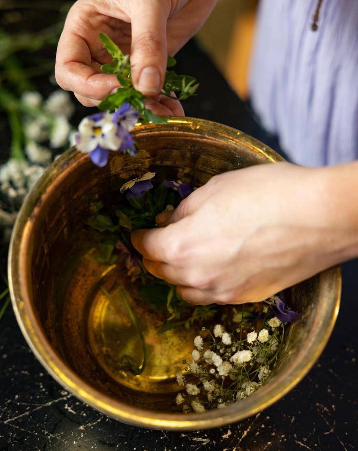 A hand from a person in a purple dress, placing flowers in a ring in a medium sized bowl
