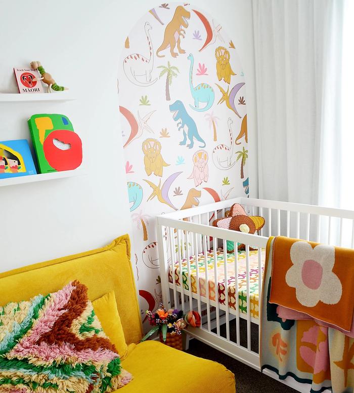 A nursery with a cot, ,a dinosaur decal in an arch above the cot, and a mustard yellow couch next to it