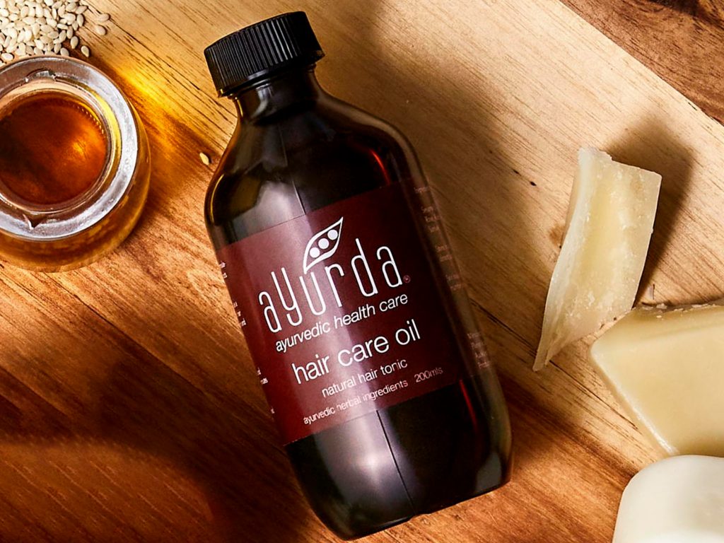 Ayurda Natural Hair Tonic lying on a wood board surrounded by natural ingredients and oils
