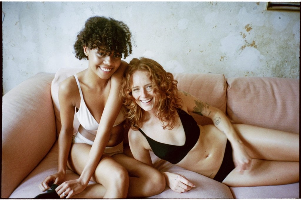 Two women sitting together on a pink couch laughing while wearing sustainable undergarments