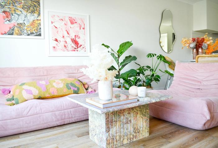 A lounge with a coffee table, a pink couch, matching chair, artwork and a mirror on the wall.