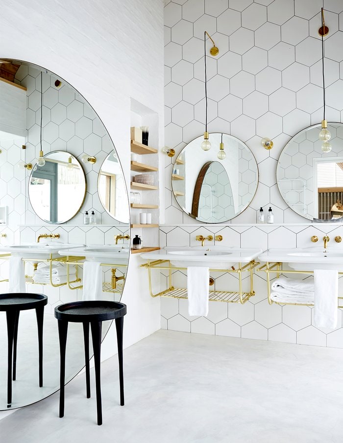 White tiled bathroom with large round mirrors, gold touches and black chair
