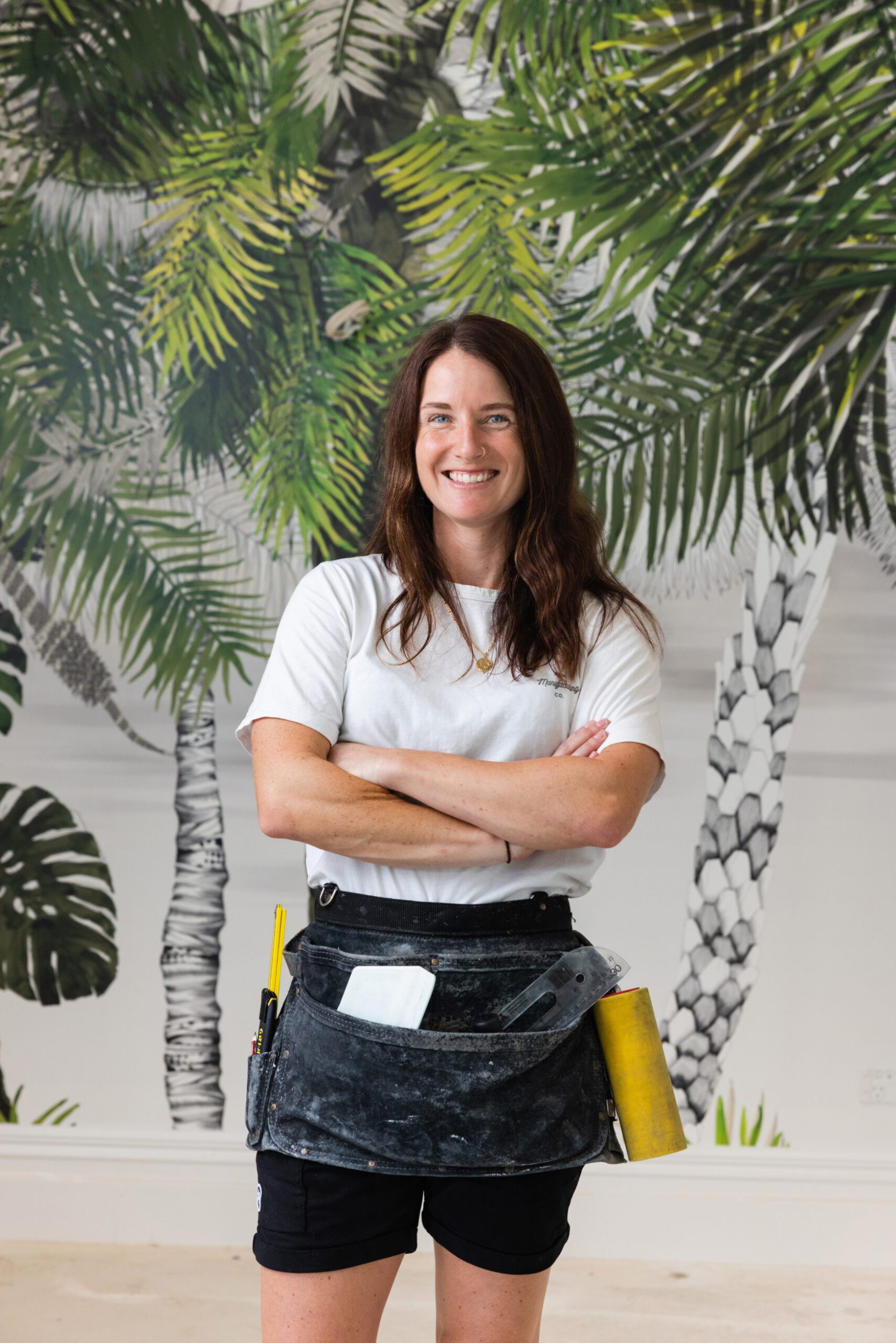 Amy Howell wearing white top and a tool belt standing against botanical themed wallpaper 