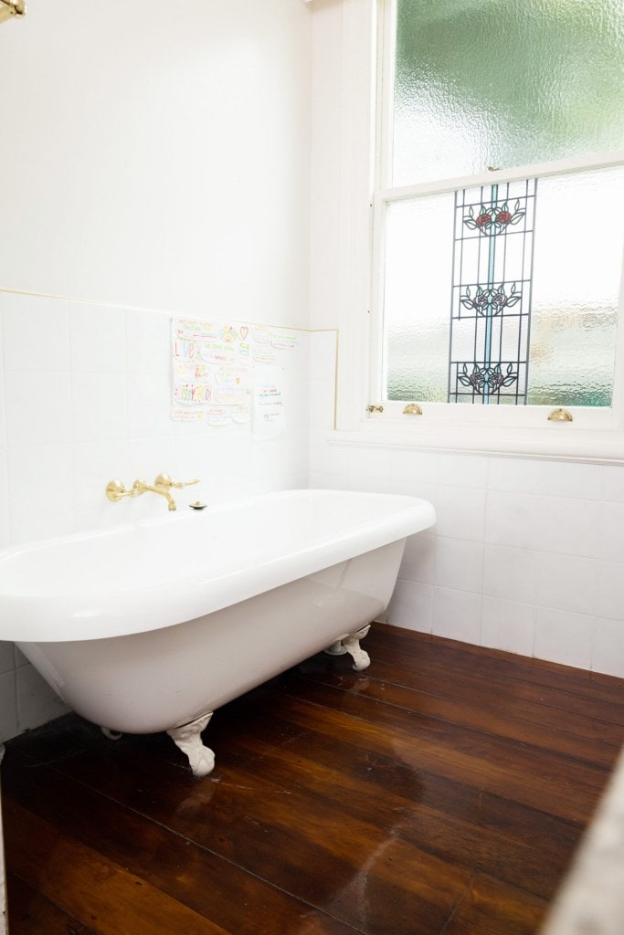 A bathroom with wood floors, white walls and a vintage grey and white tub with gold taps