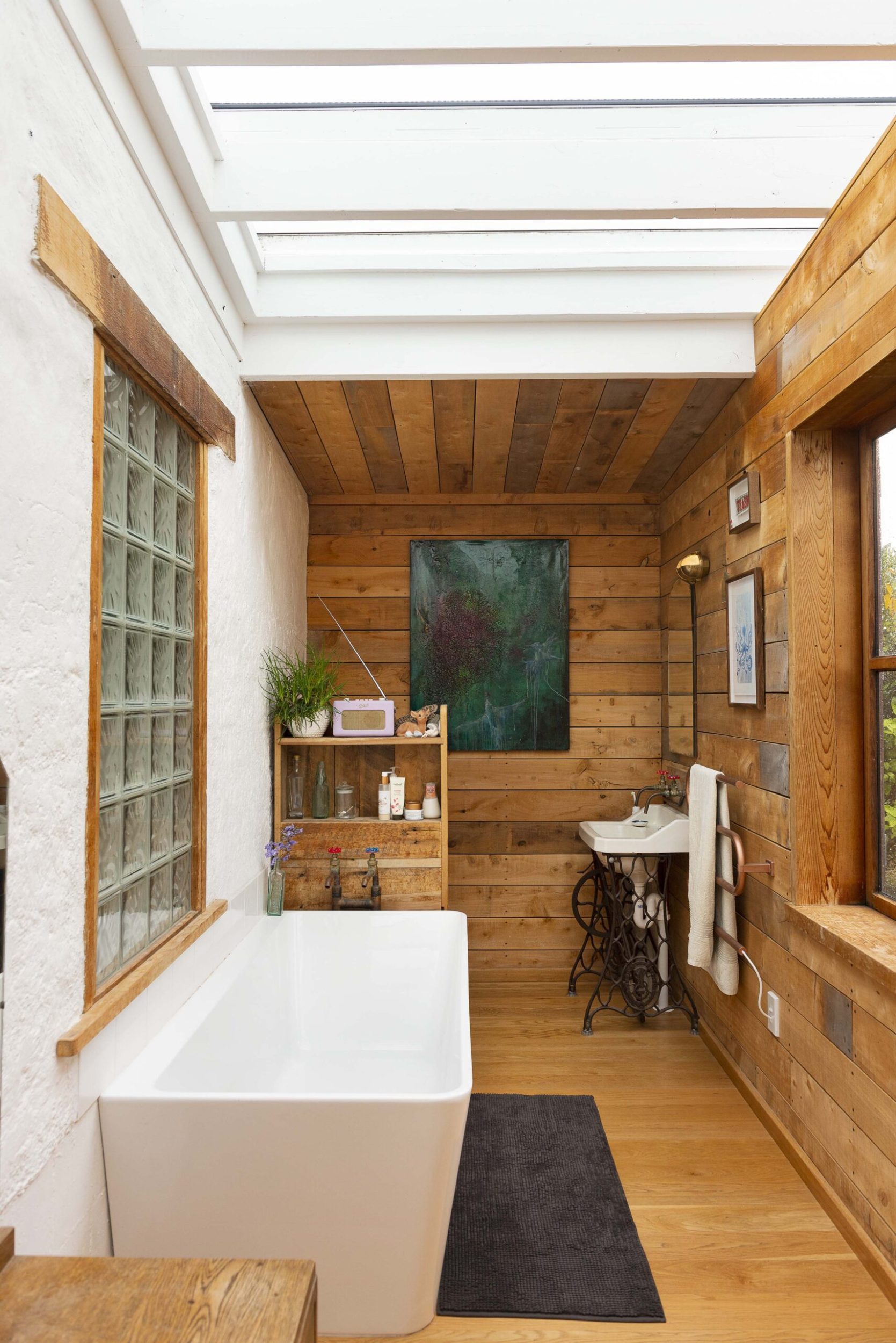 A bathroom with white and wood panelled walls, a large white bathtub, ceiling light and hanging blue art