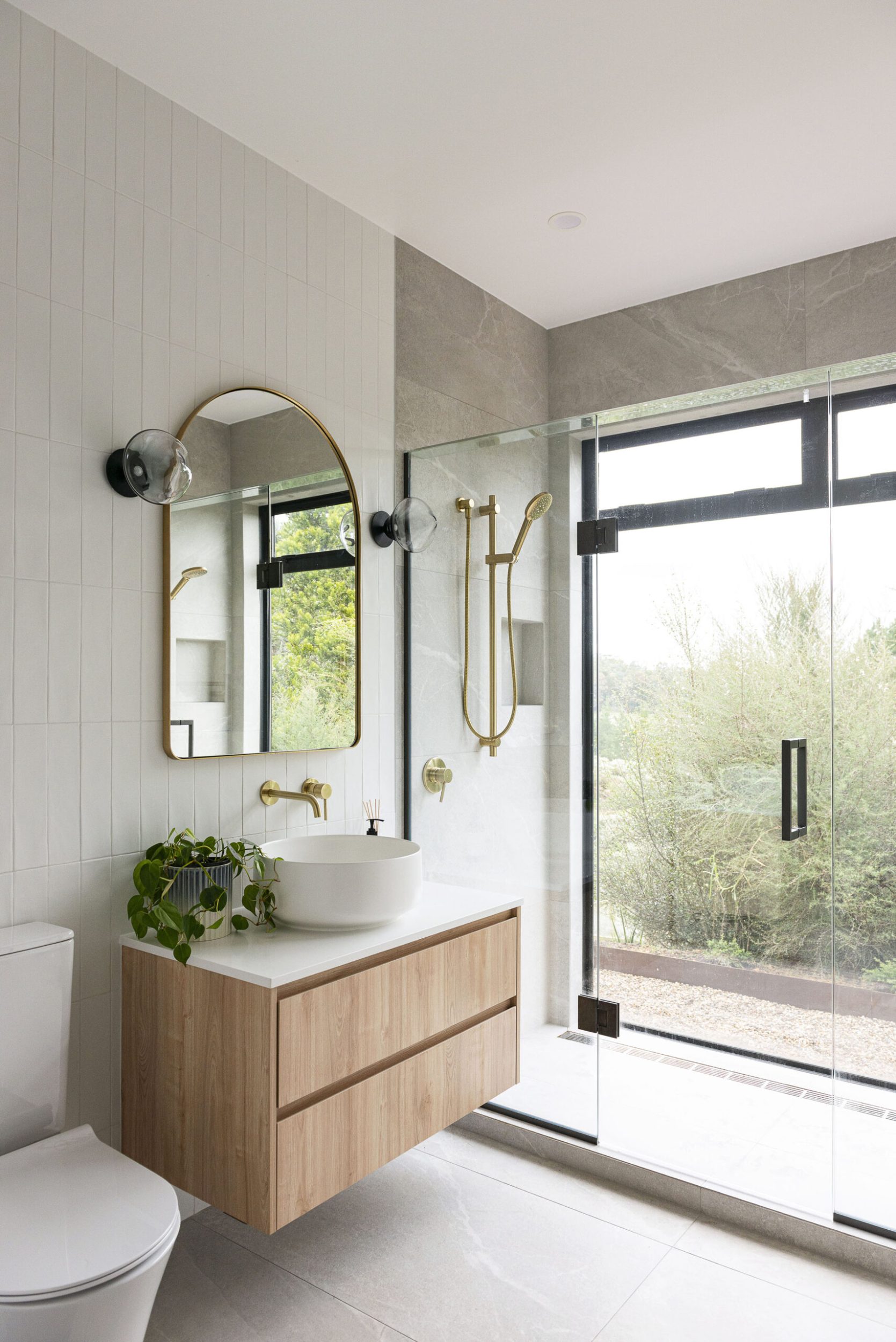 A bathroom with grey tiles, a wood hanging cabinet, gold framed mirror and large open window 