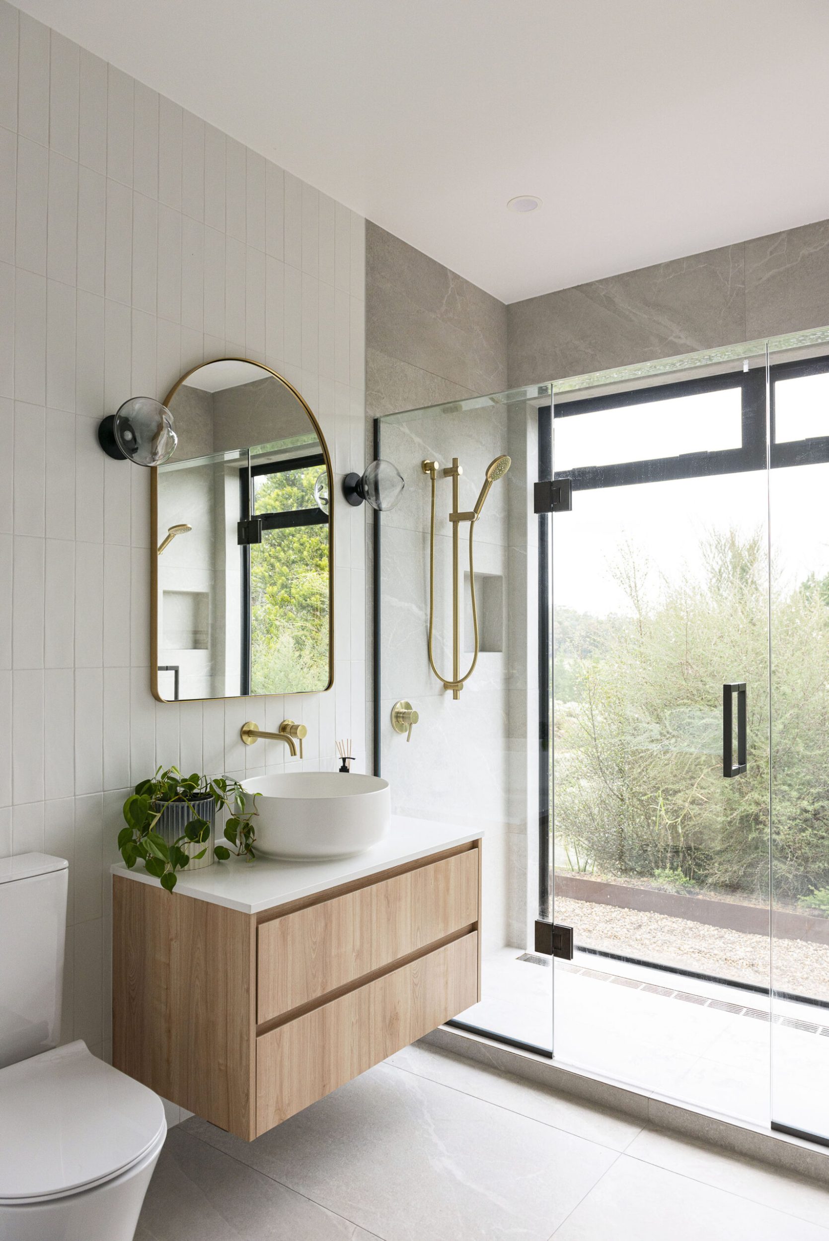 A bathroom with grey tiles, a wood hanging cabinet, gold framed mirror and large open window