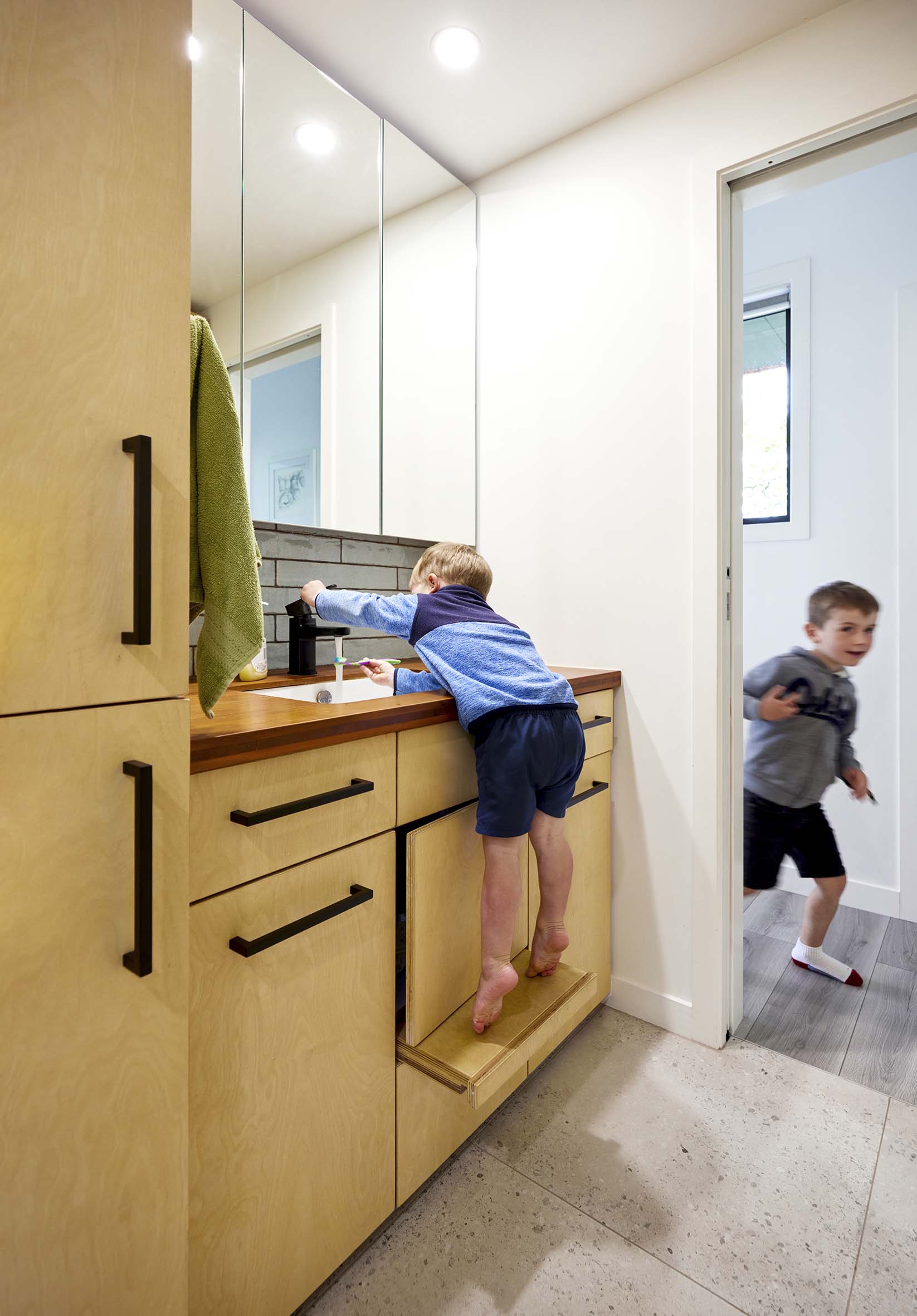 Two young boys in a bathroom, with one of them leaning to turn on sink while standing on a in-build stand