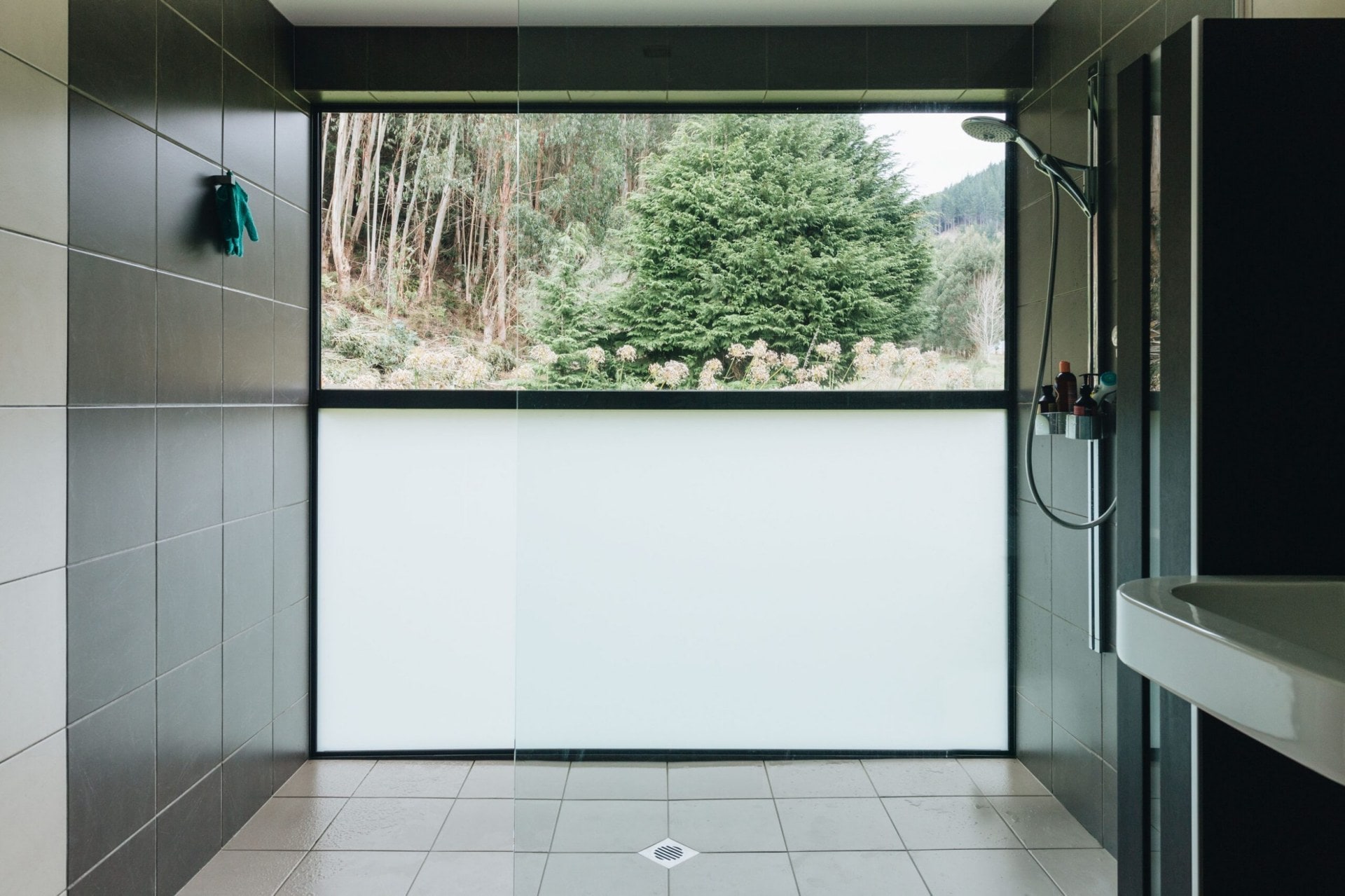 A large shower with black wall tiles, grey floor tiles and large windows facing bushland 