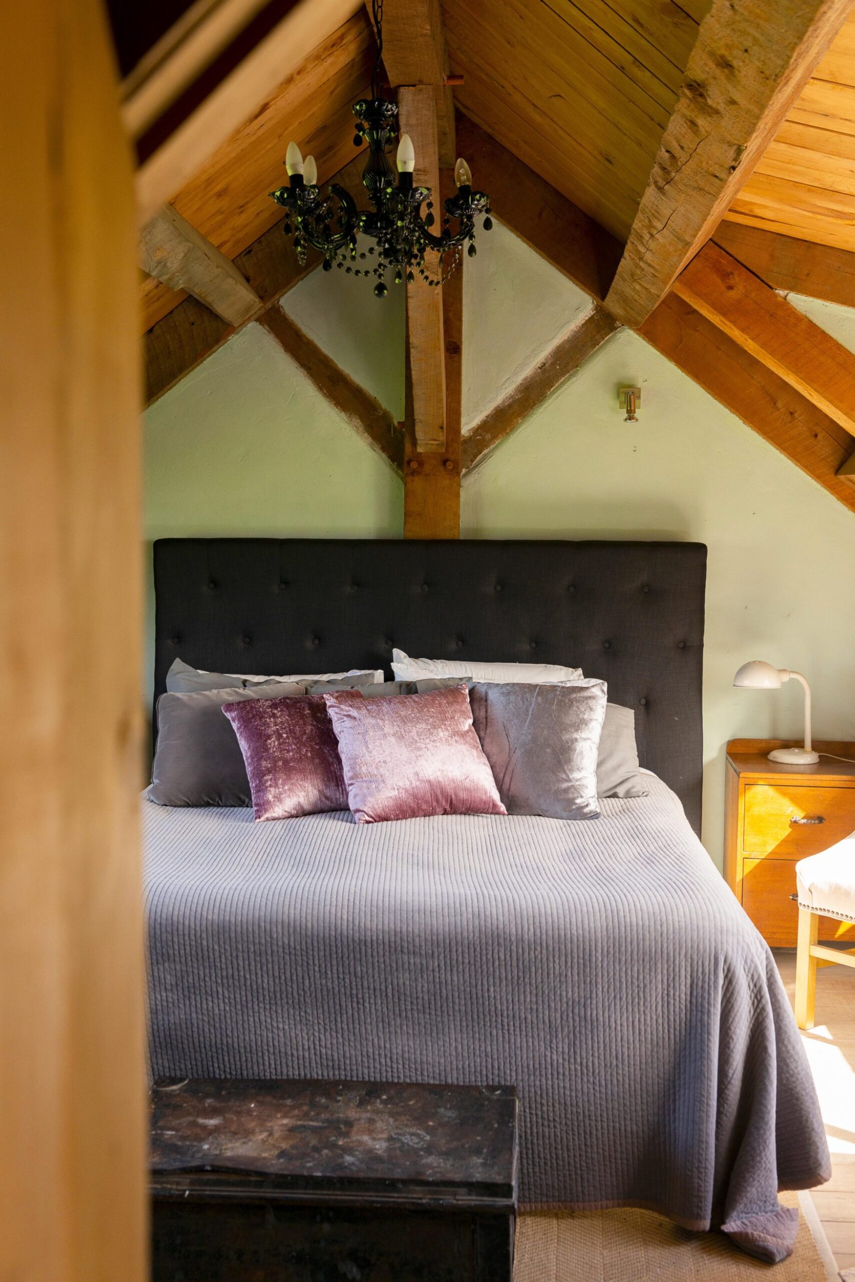 A bedroom with limewashed green walls, a large bed with a black bedboard and arched wooden ceilings 