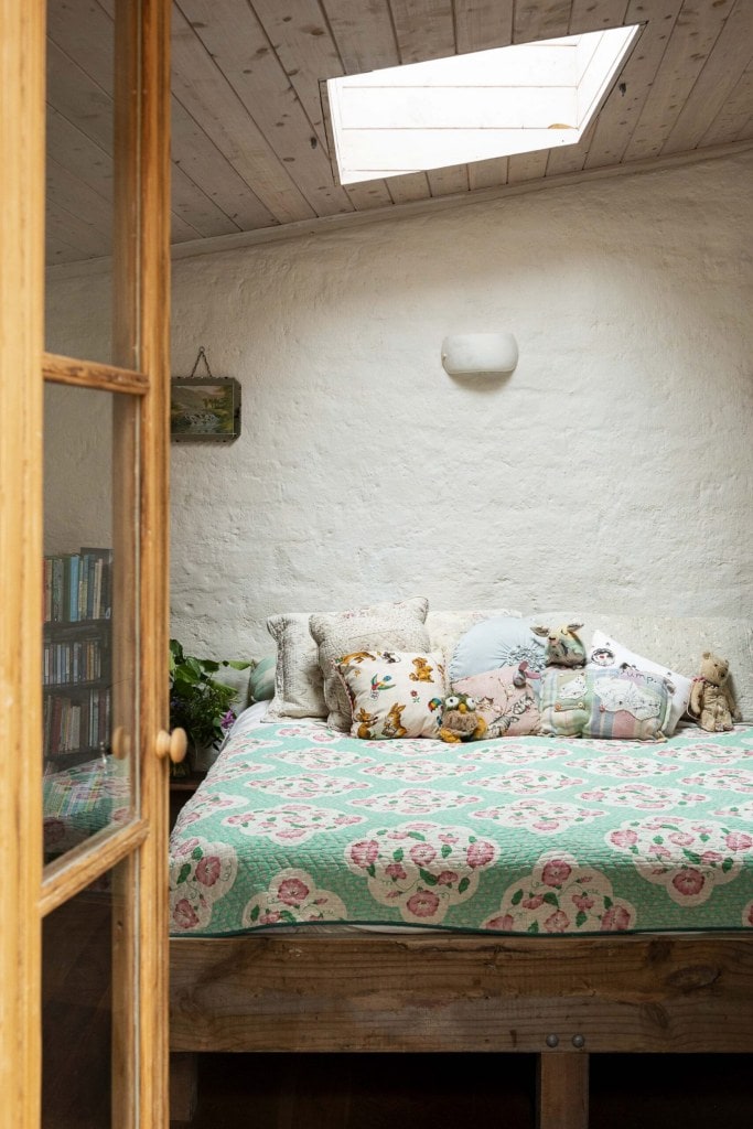 A bedroom with white walls, light wood ceiling, a ceiling light, and a colourful teal duvet and cushions