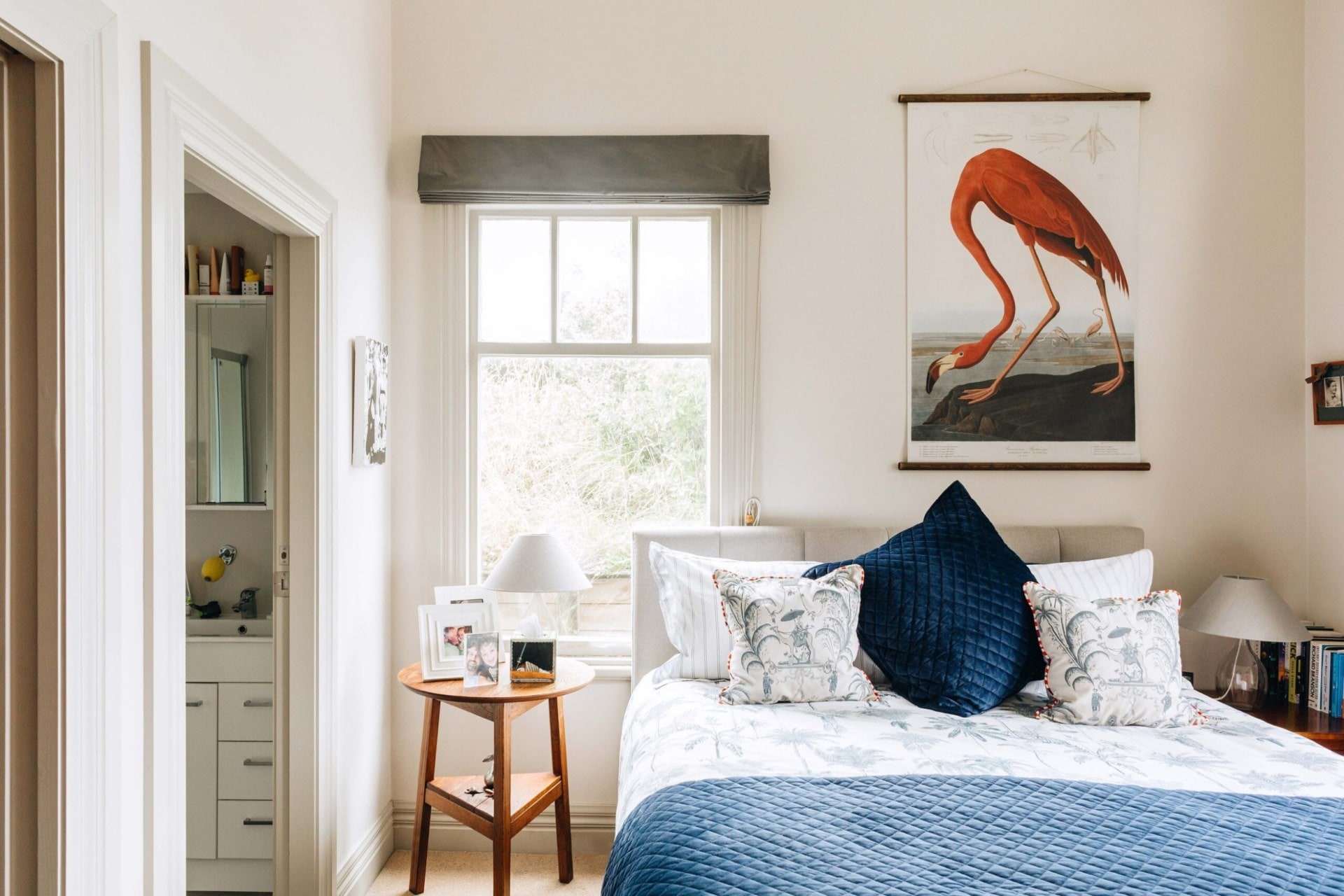 A bedroom with a bed with a blue and white duvet, a round wood side table and a large hanging art print of a flamingo