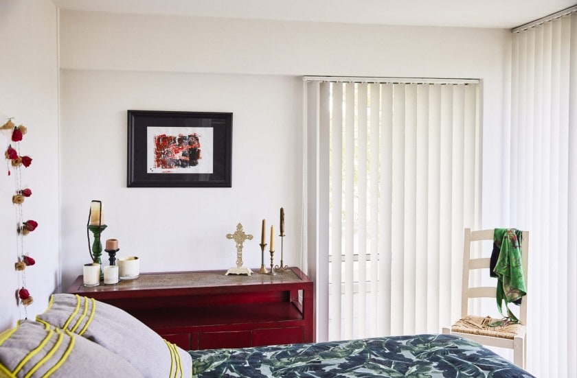 White bedroom with a bed, hanging art and a red Asian-style console in the corner