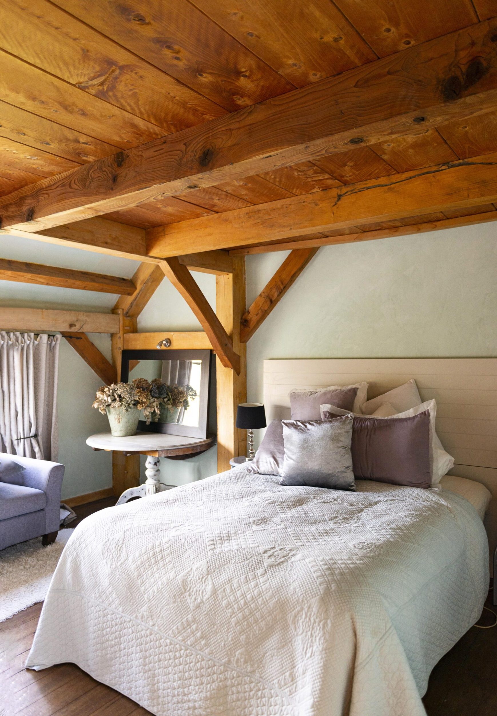 A bedroom in a cottage home with wood beams, a bed with a white duvet and limewashed green walls