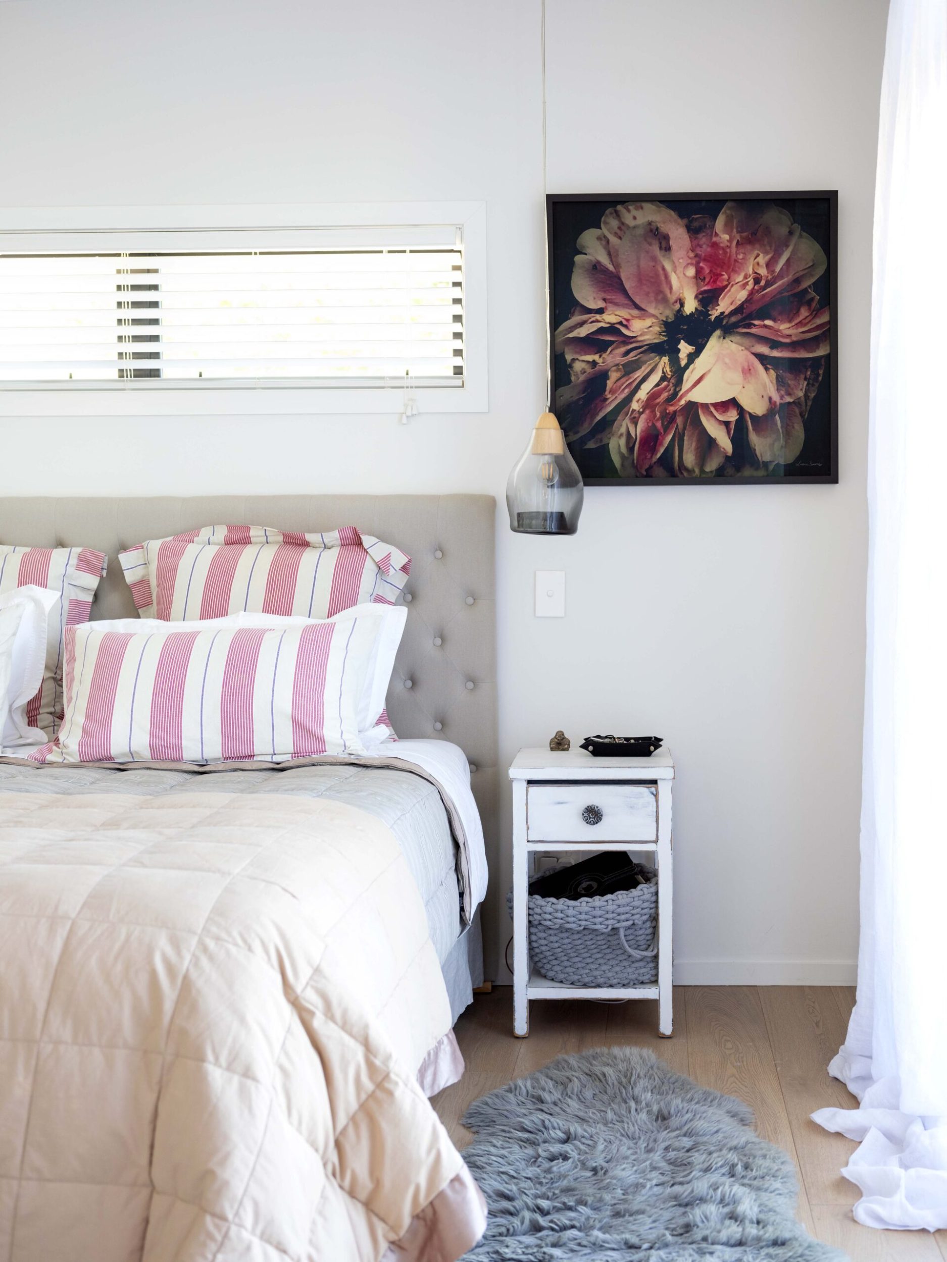 A white bedroom with a bed with pink and white striped pillows and hanging art by Lizzie Beere