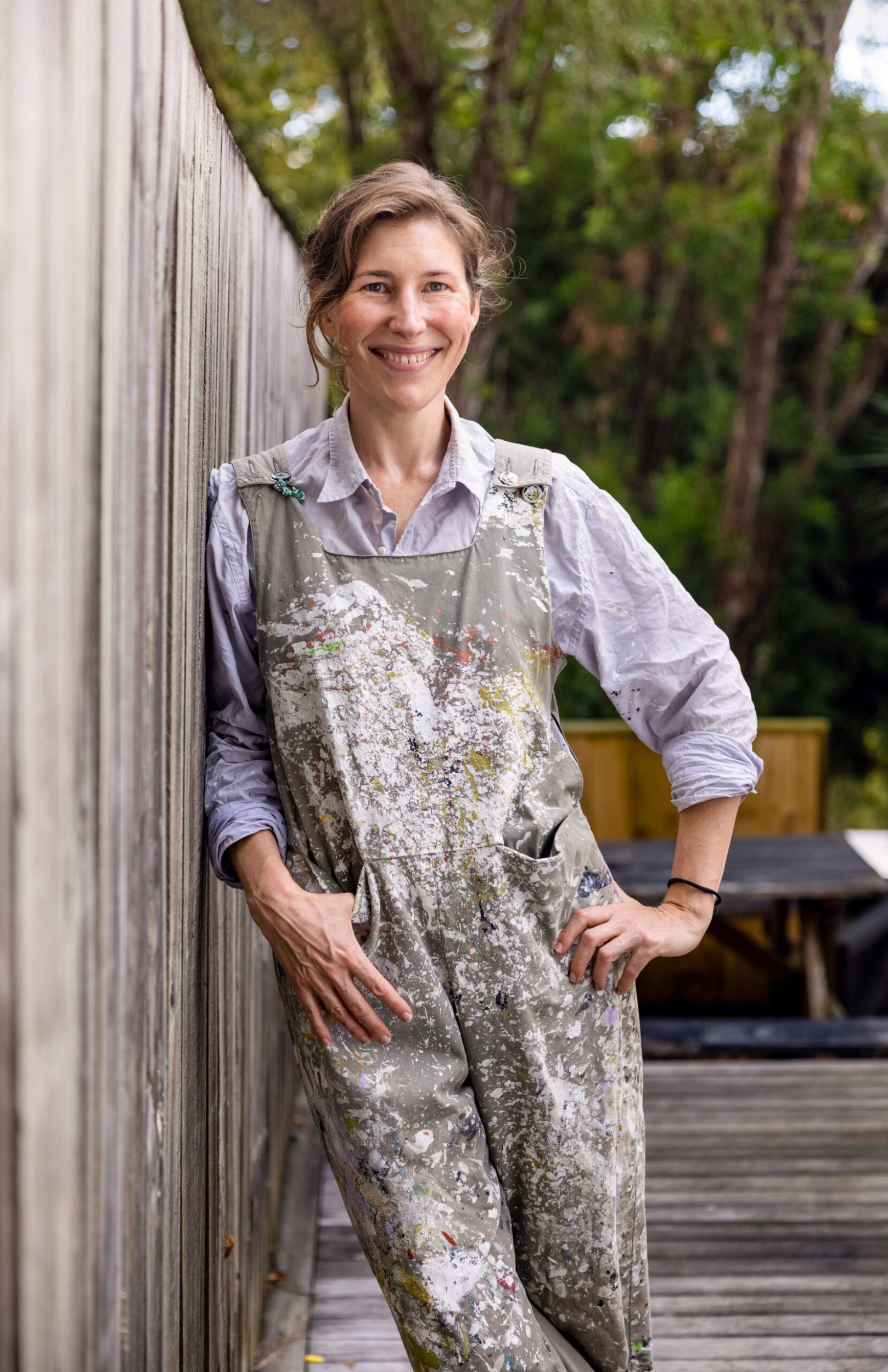 Beth Ellery smiling while leaning against fence wearing paint splattered overalls 