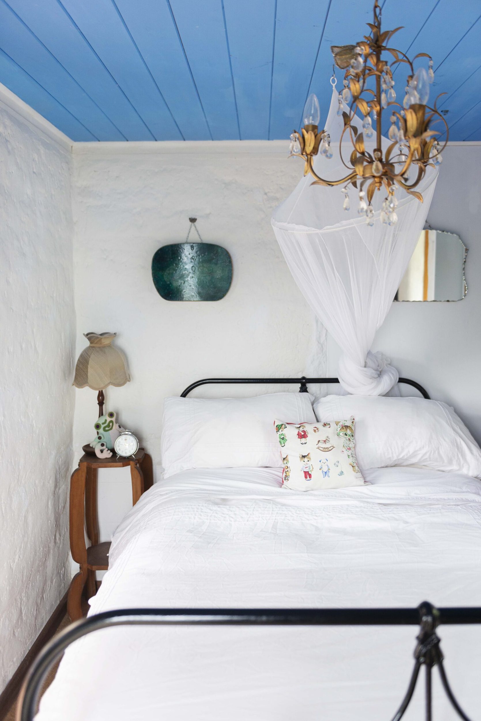 A bedroom with white walls, blue panel ceilings, a gold chandelier and a bed with a black frame