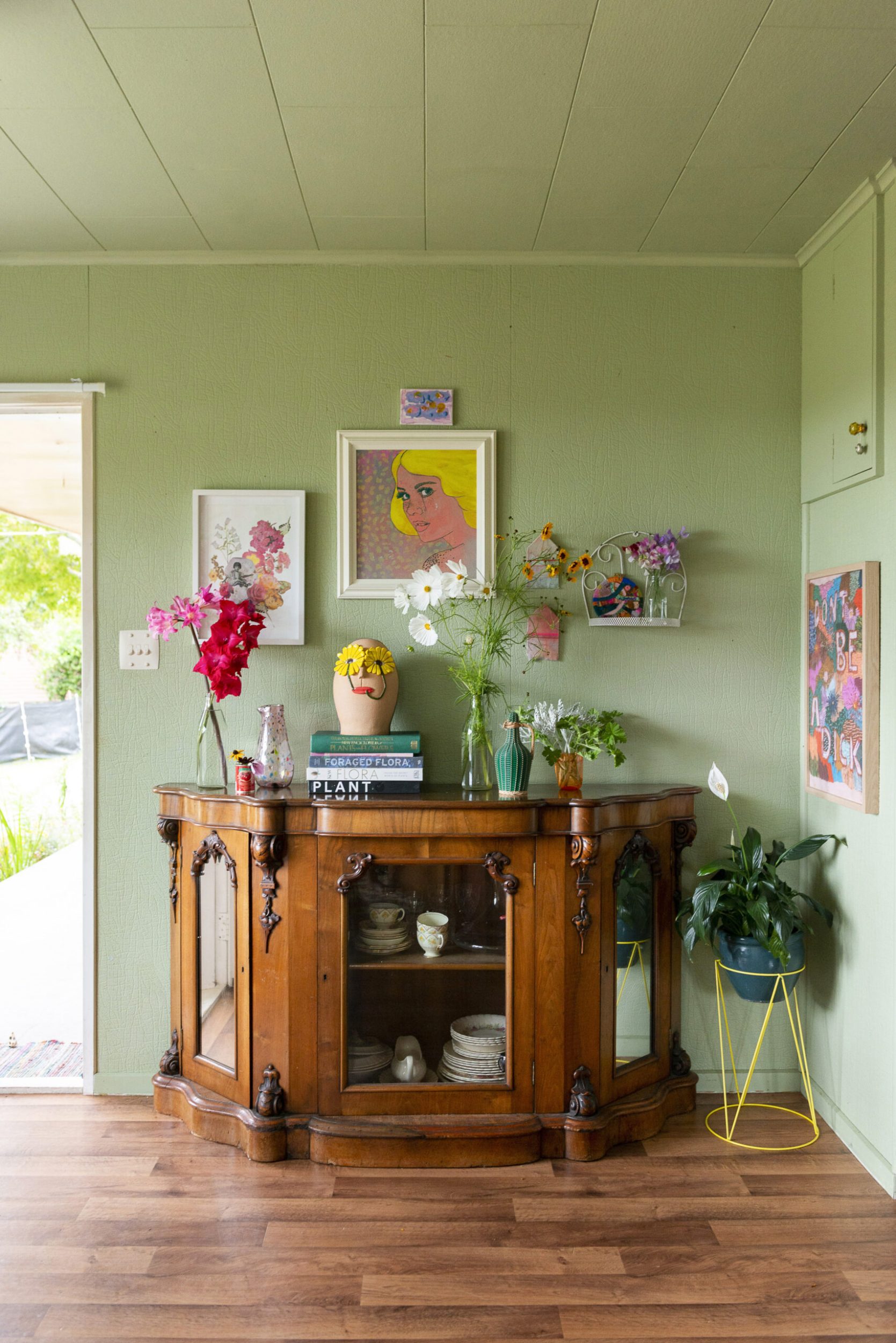 Green room with wood floors and a brown vintage cabinet stacked with books and vases filled with flowers 