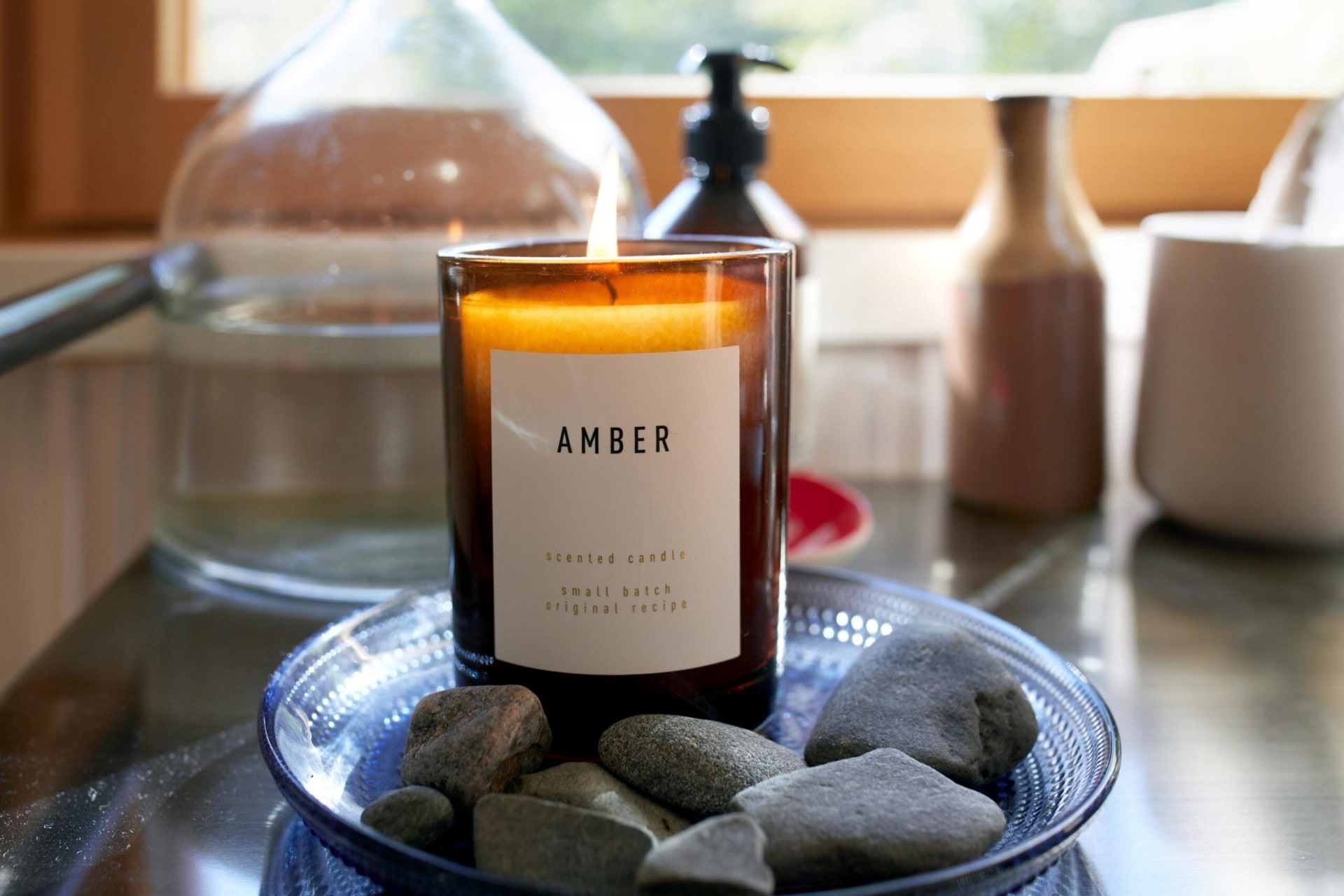 A lit amber coloured Amber brand candle set in a round blue plate next to grey rocks