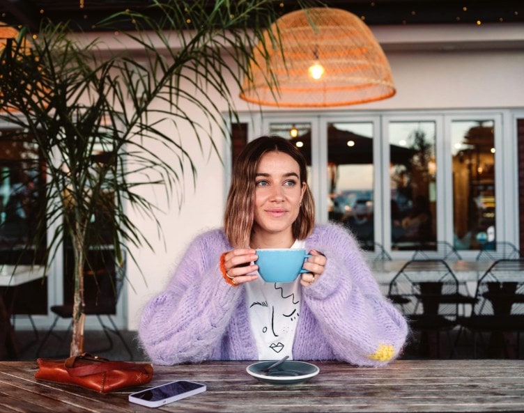 Danni Duncan sitting outside a cafe drinking coffee while wearing a lavender cardigan