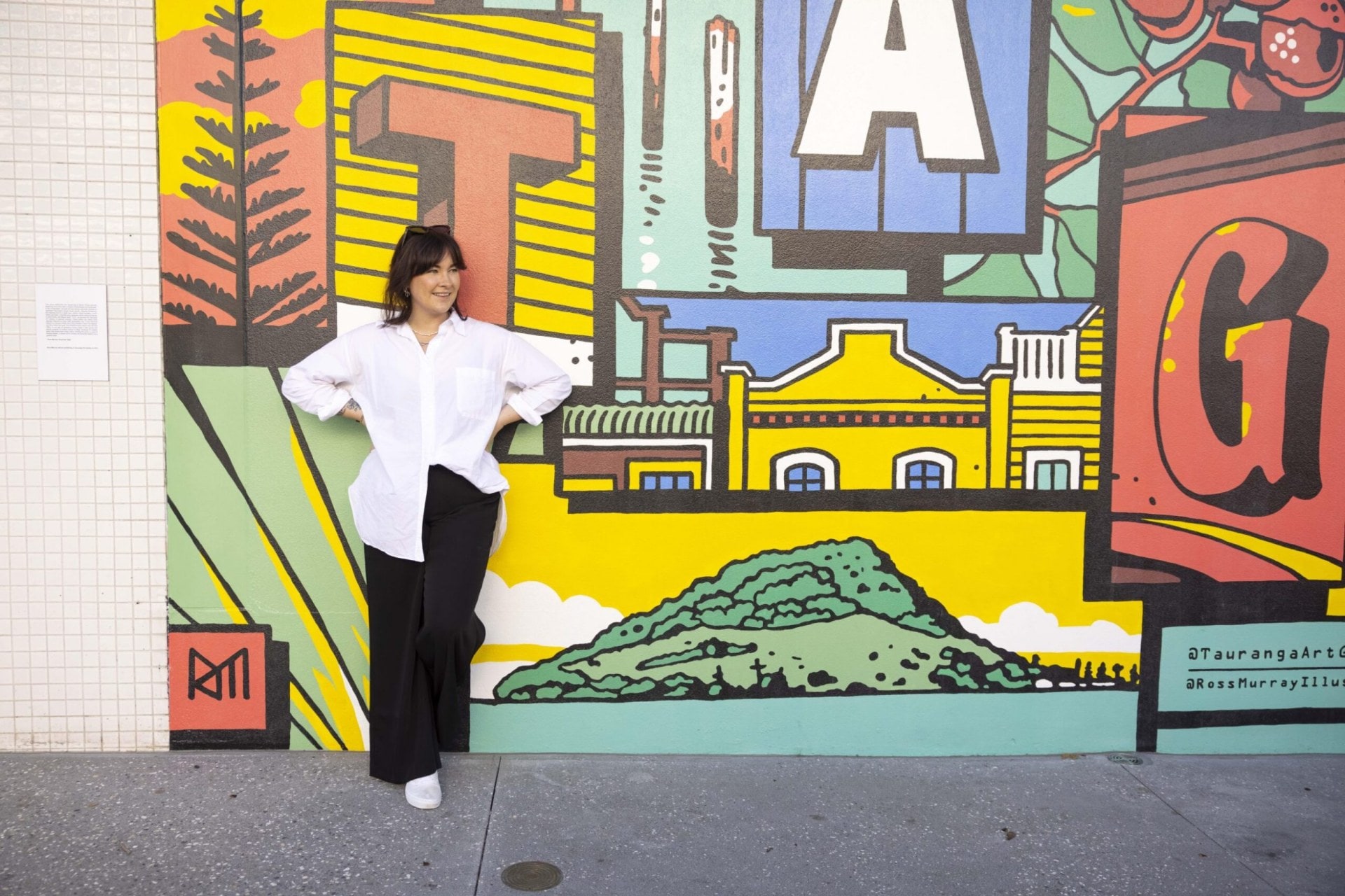 Lucy Burke leaning against a colourful Mount Maunganui mural by Ross Murray outside of Tauranga Art Gallery