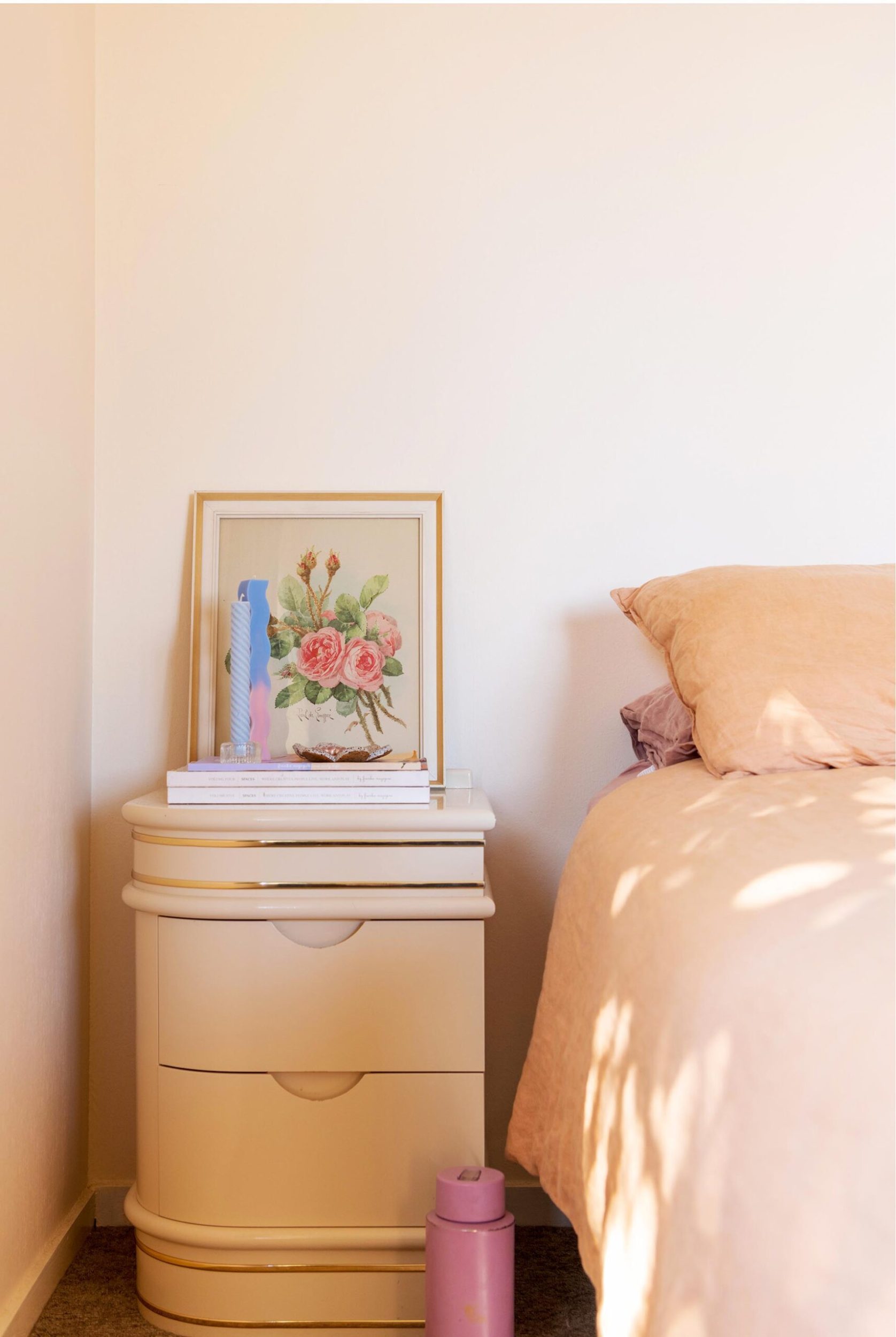 A peach coloured bedroom with a curved bedside table with a floral artwork resting on top 