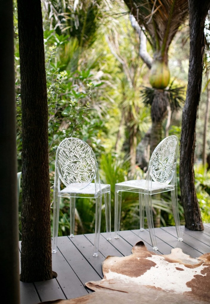 A black deck set by green bush and with two clear plastic chairs on it