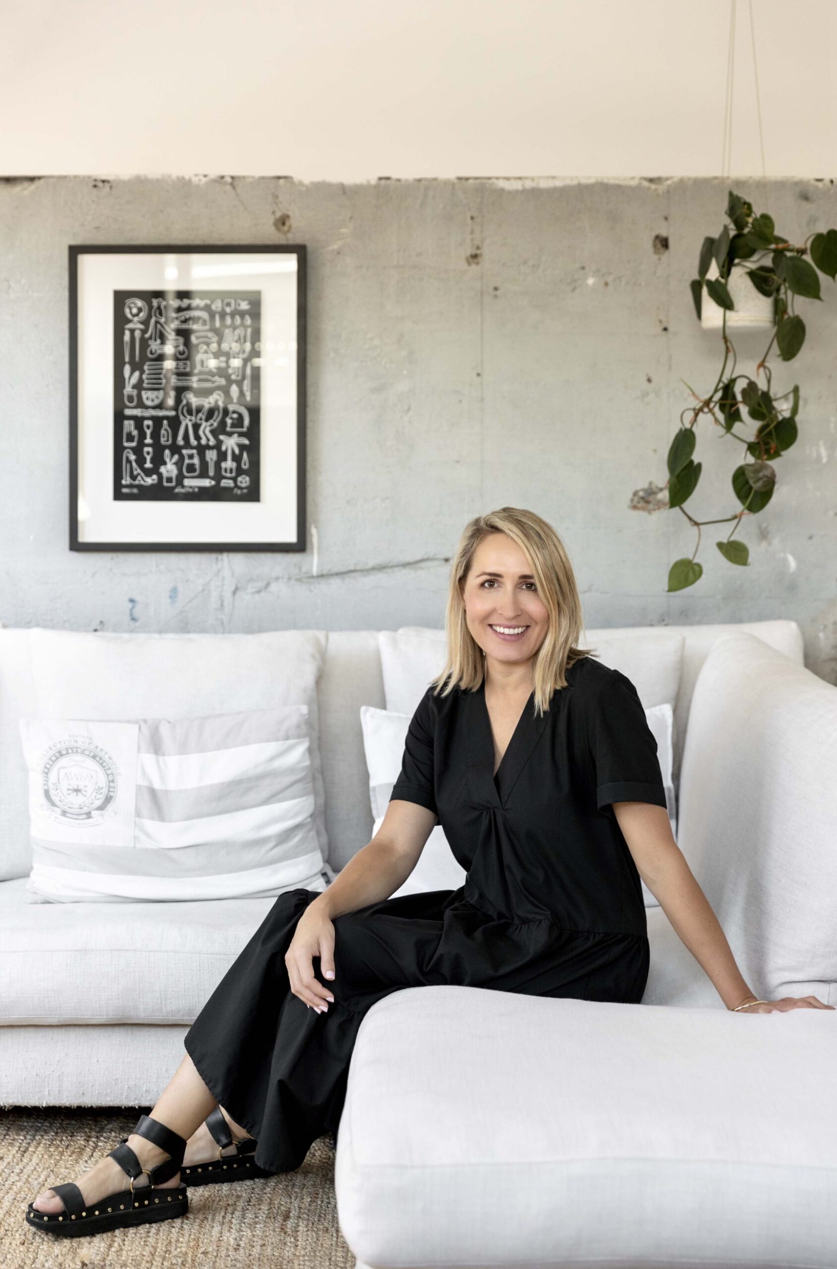 Dee Johnson sitting on a white couch wearing a black jumpsuit with art by Andrew J Steel hanging on a wall 