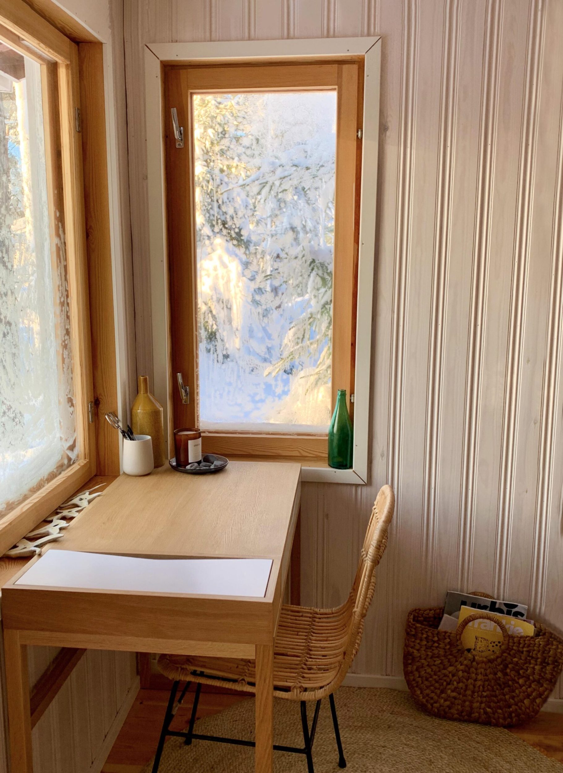 A light brown wood desk set in the corner of a cabin with snow out the window