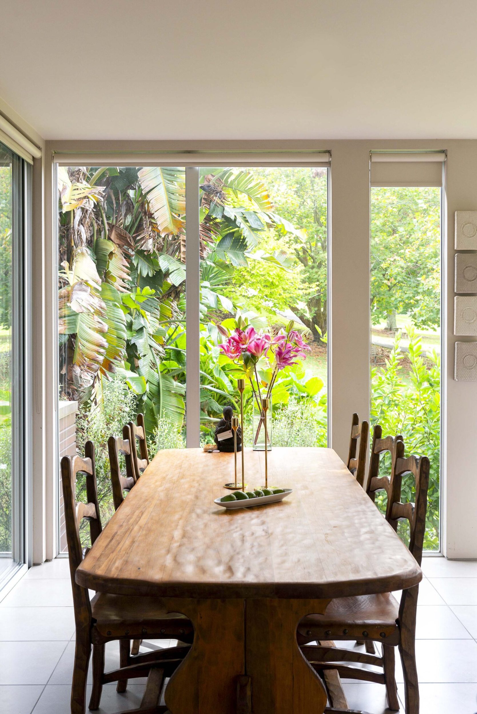 A brown dining table in a room with grey tiles and tall windows showing leafy backyard