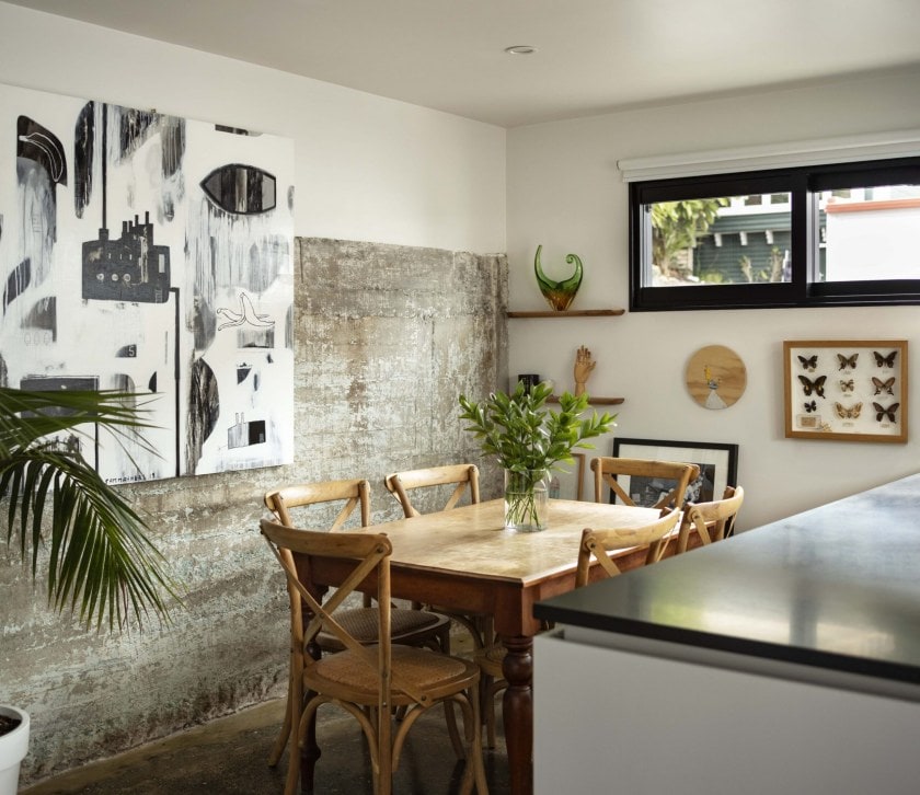A dining room with a wall with a white and exposed concrete wall, a brown dining table and green and brown hanging art 