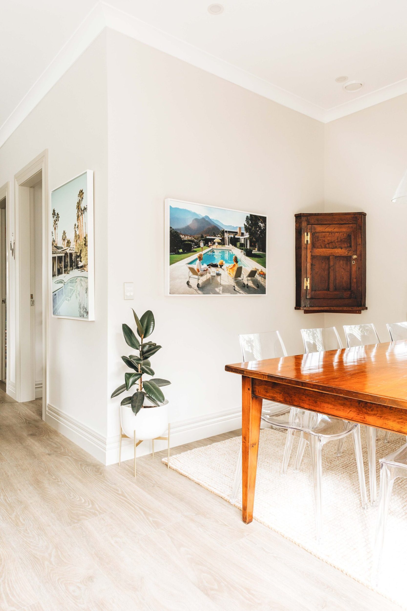 A white dining room with a green plant, a brown shelf and a Slim Aarons Palm Springs hanging photo