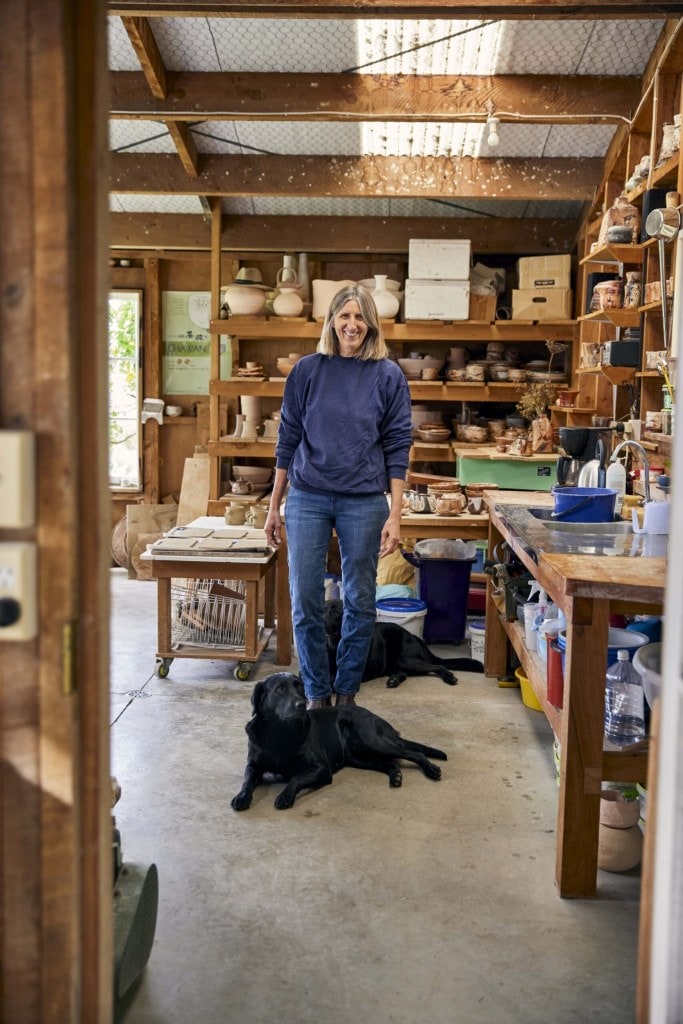 Elena Renker standing in her art studio surrounded by ceramic bowls with her black dogs dogs Mika and Hanna at her feet